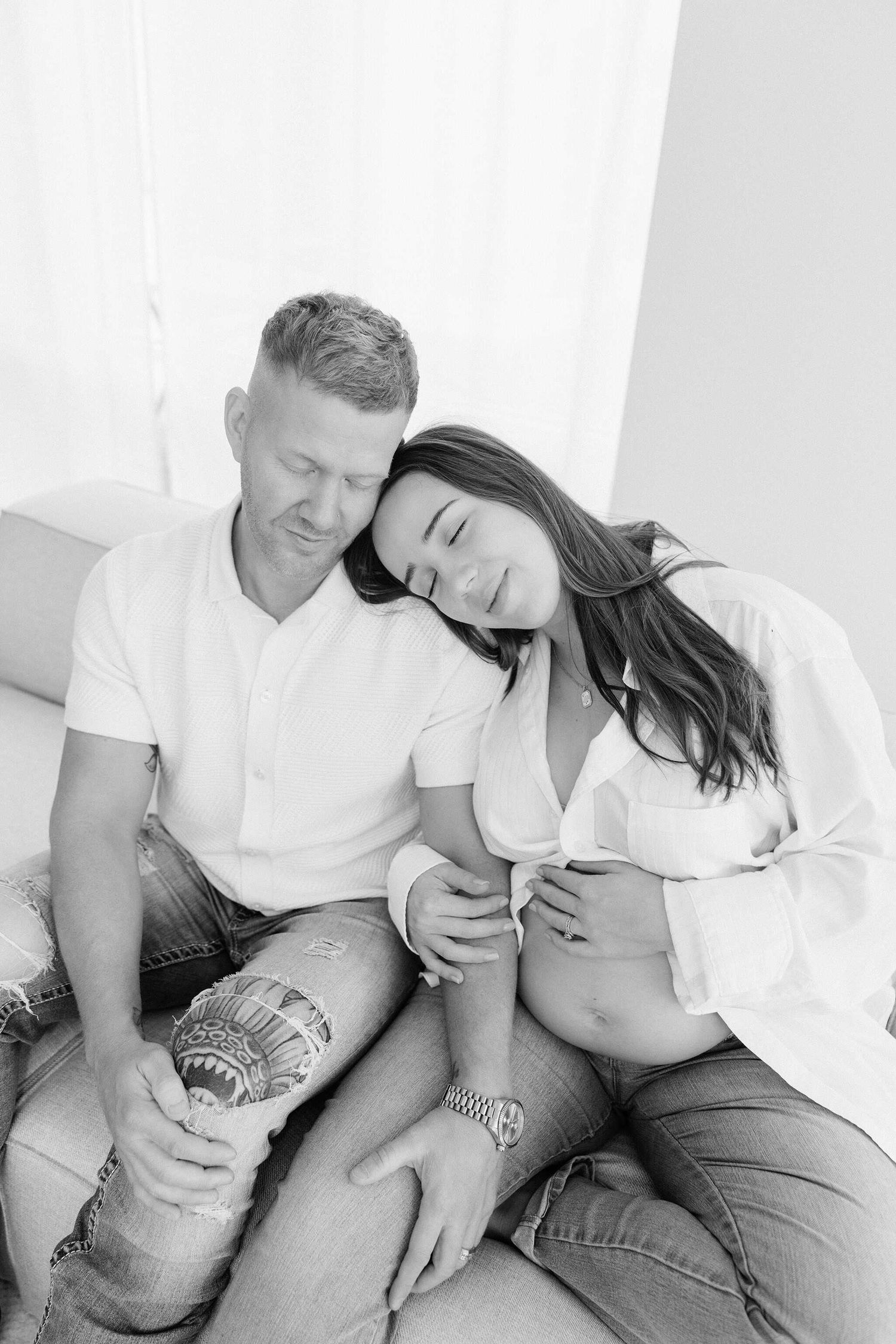 black and white photo of maternity couple sitting on the couch with mom leaning onto dads shoulder