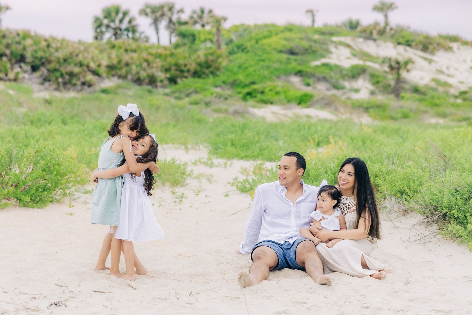mom and dad holding baby and smiling at 2 daughters hugging on the beach