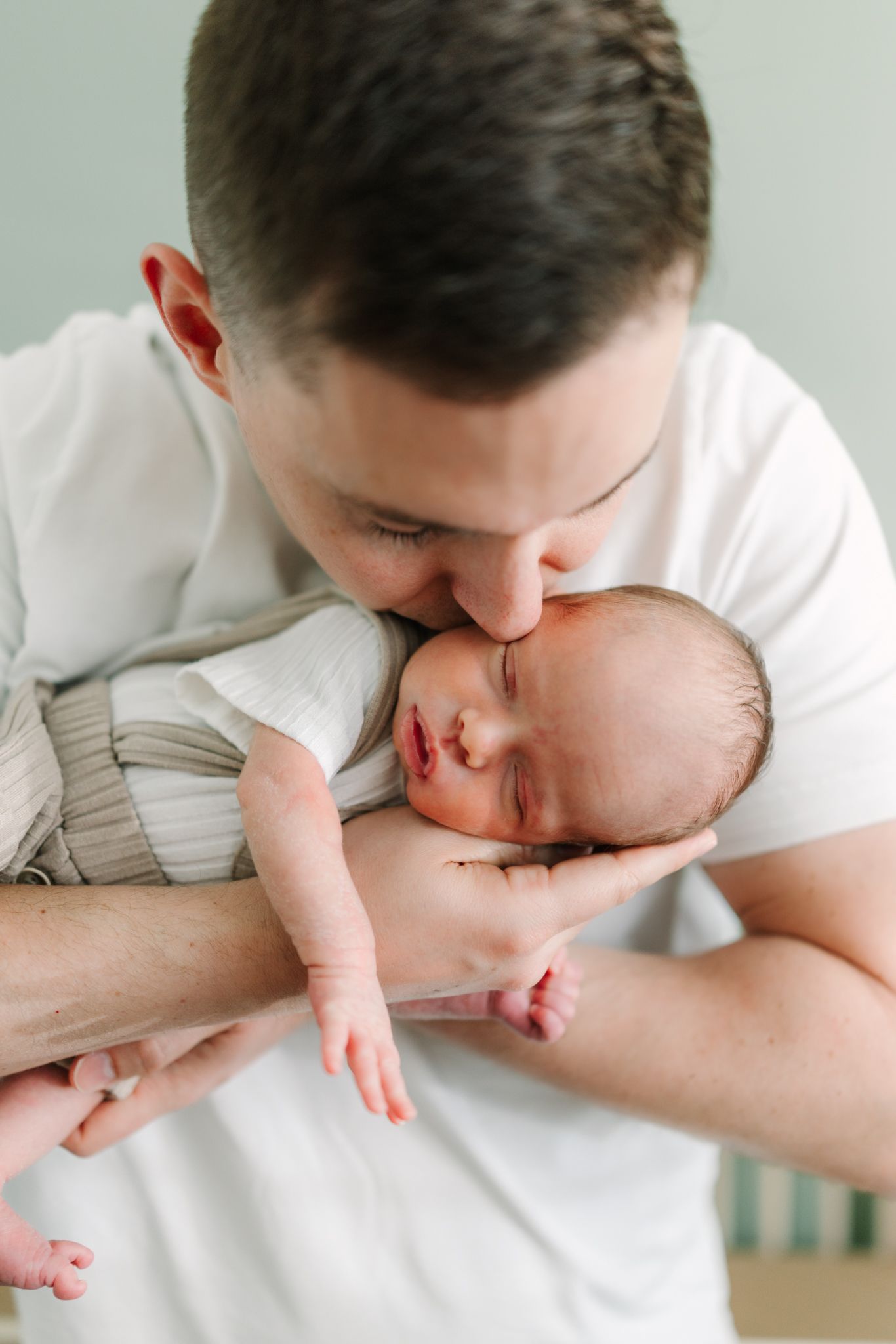 A father in a white shirt gently kisses his sleeping newborn baby cradled in his arms.
