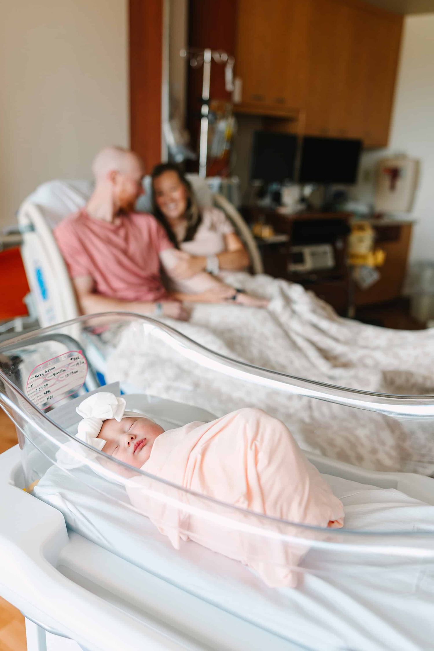 newborn baby in hospital bassinet with mom and dad sitting in hospital bed behind