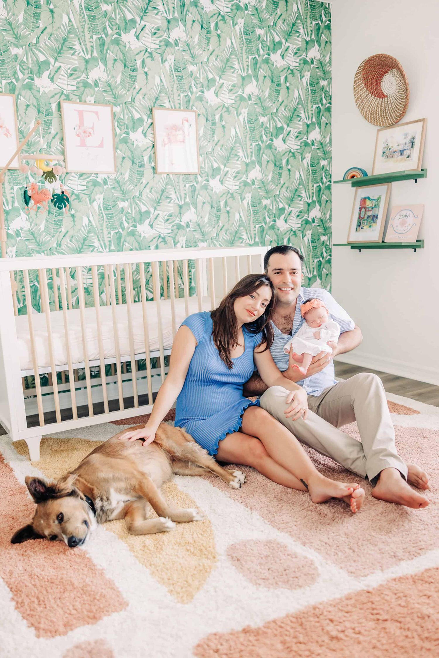 mom leaning on dads shoulder while they both sit in front of the crib with their newborn baby and mom is patting the dog