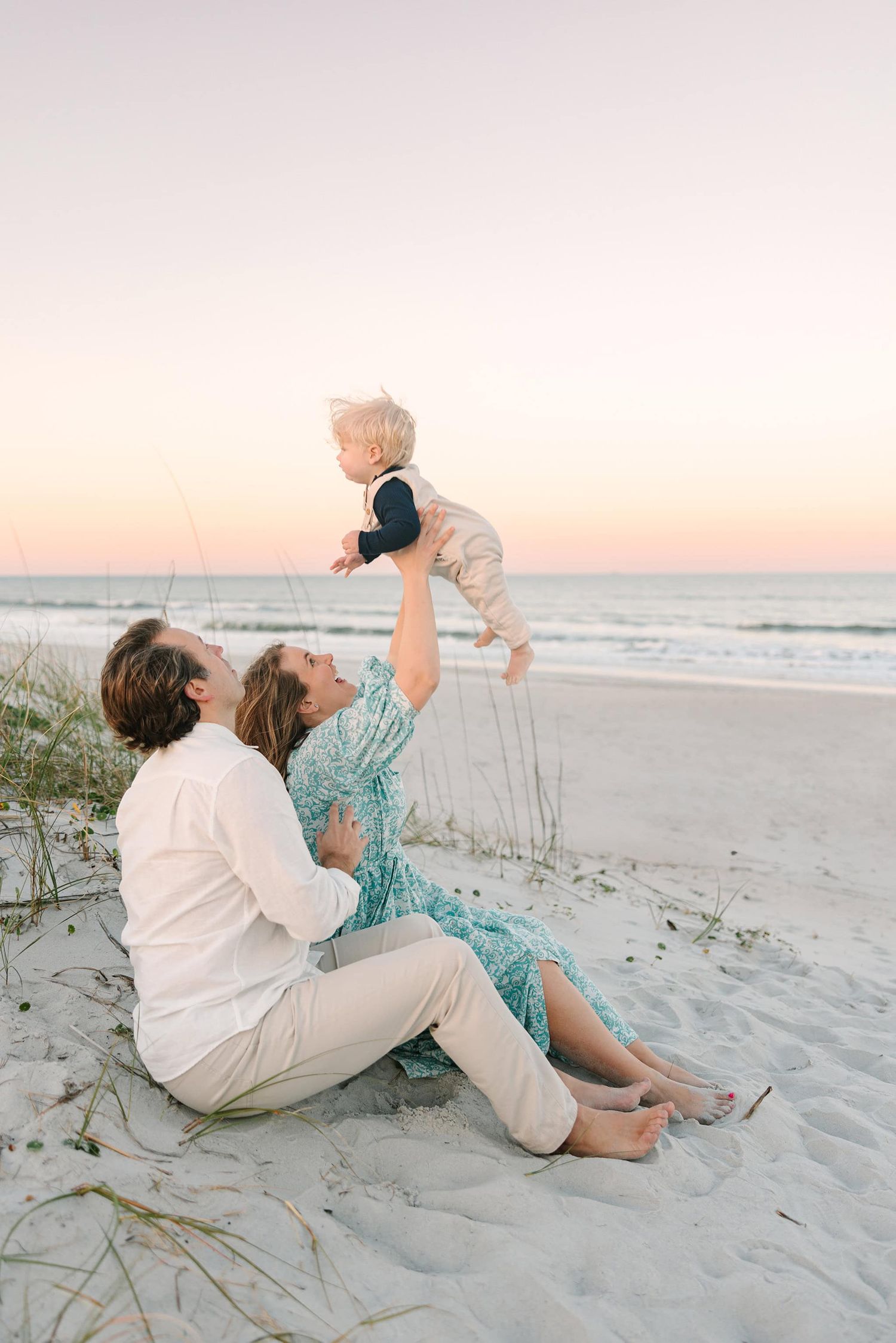 dad sitting behind mom in the sand while she throws baby boy up into the air at Jacksonville sunset