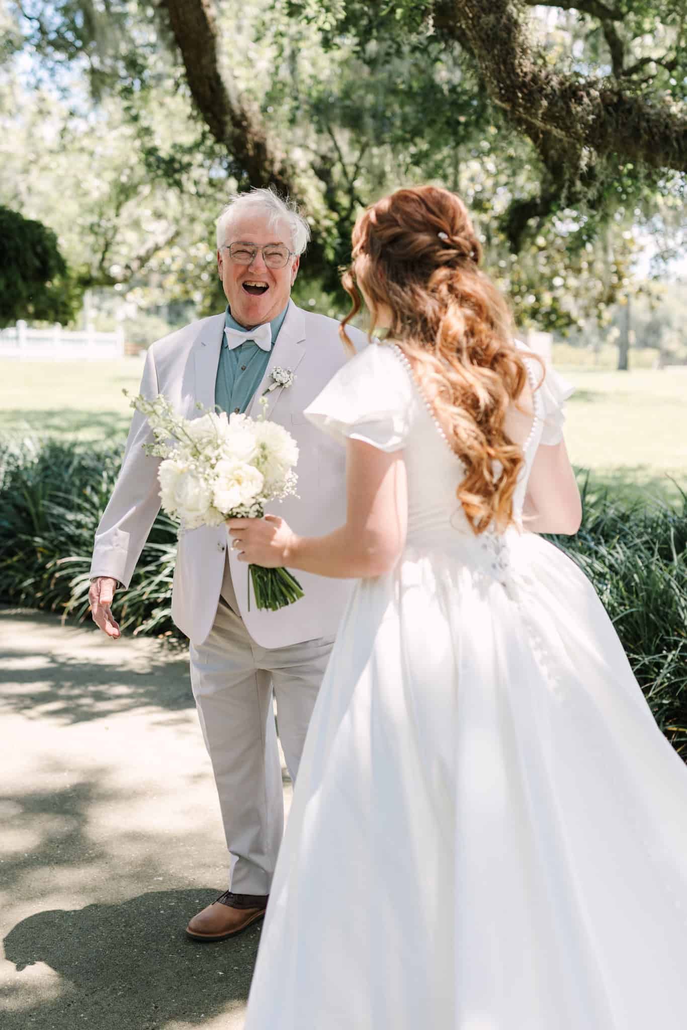 Father reacts with joy seeing bride in white wedding dress holding bouquet outdoors under oak trees.