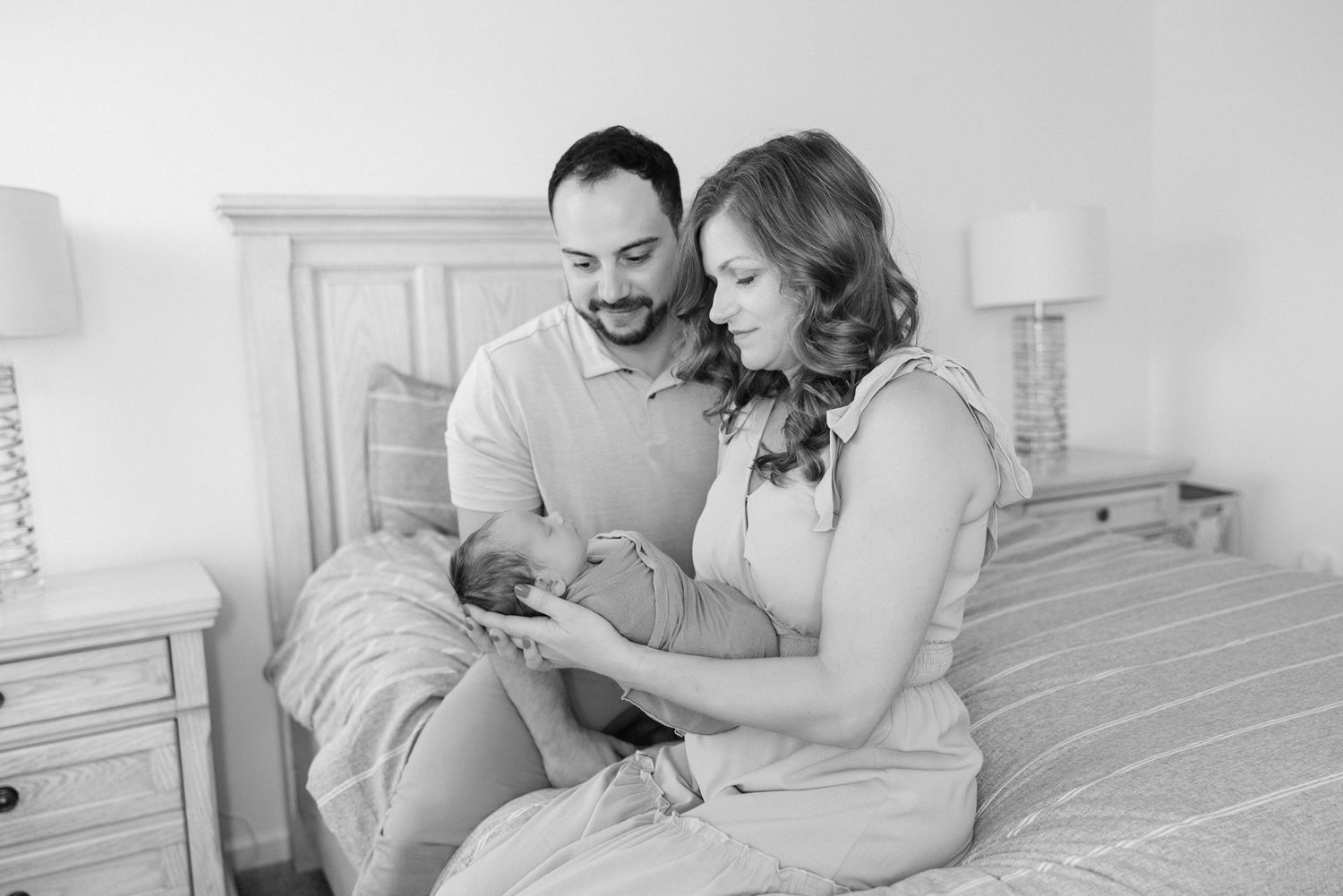 mom and dad sitting on bed holding swaddled newborn baby