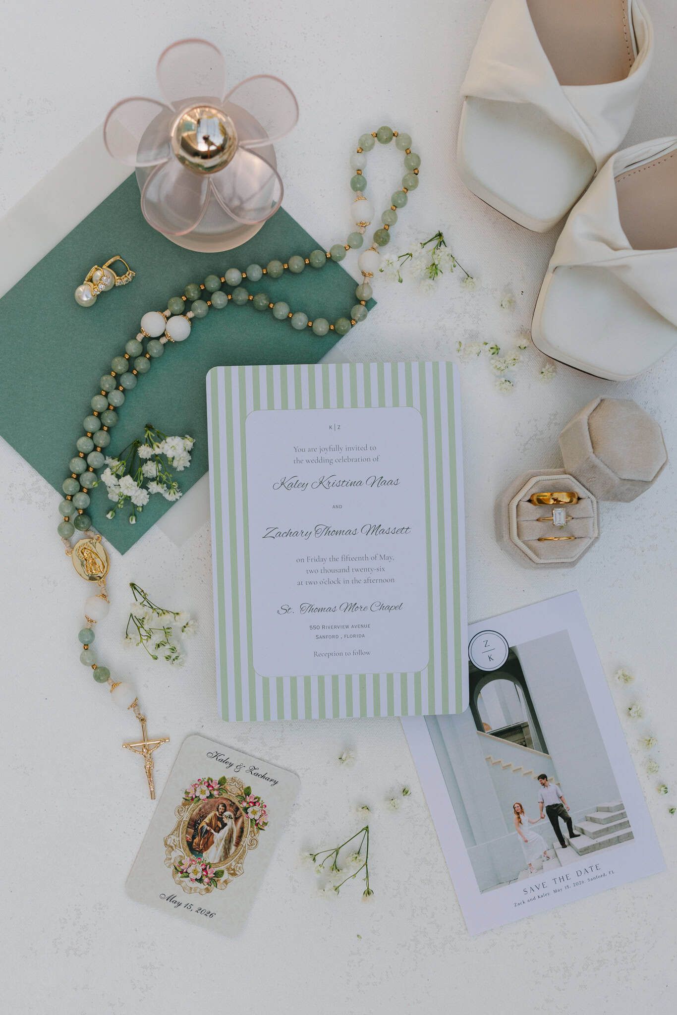 Elegant wedding flat lay with green striped invitation, rosary beads, bridal heels, perfume, and floral accents on white background.
