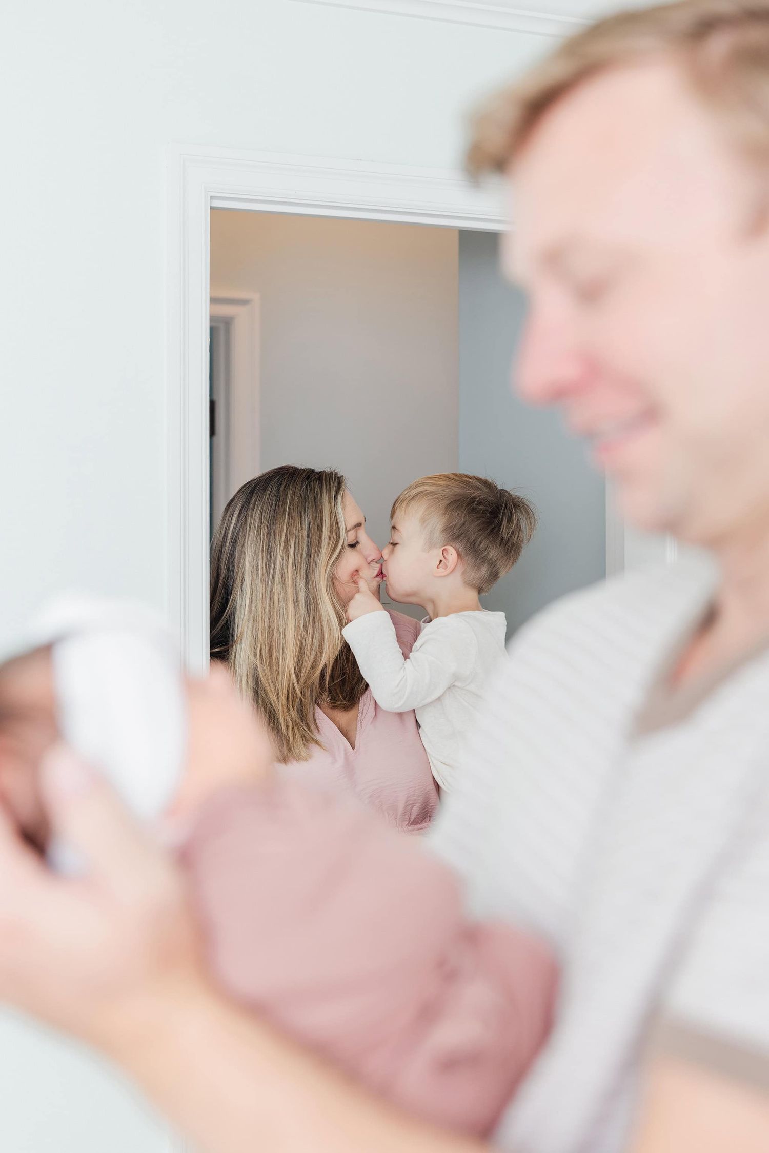 mom holding toddler son and giving him a kiss in the doorway, peeking through dad holding newborn baby girl
