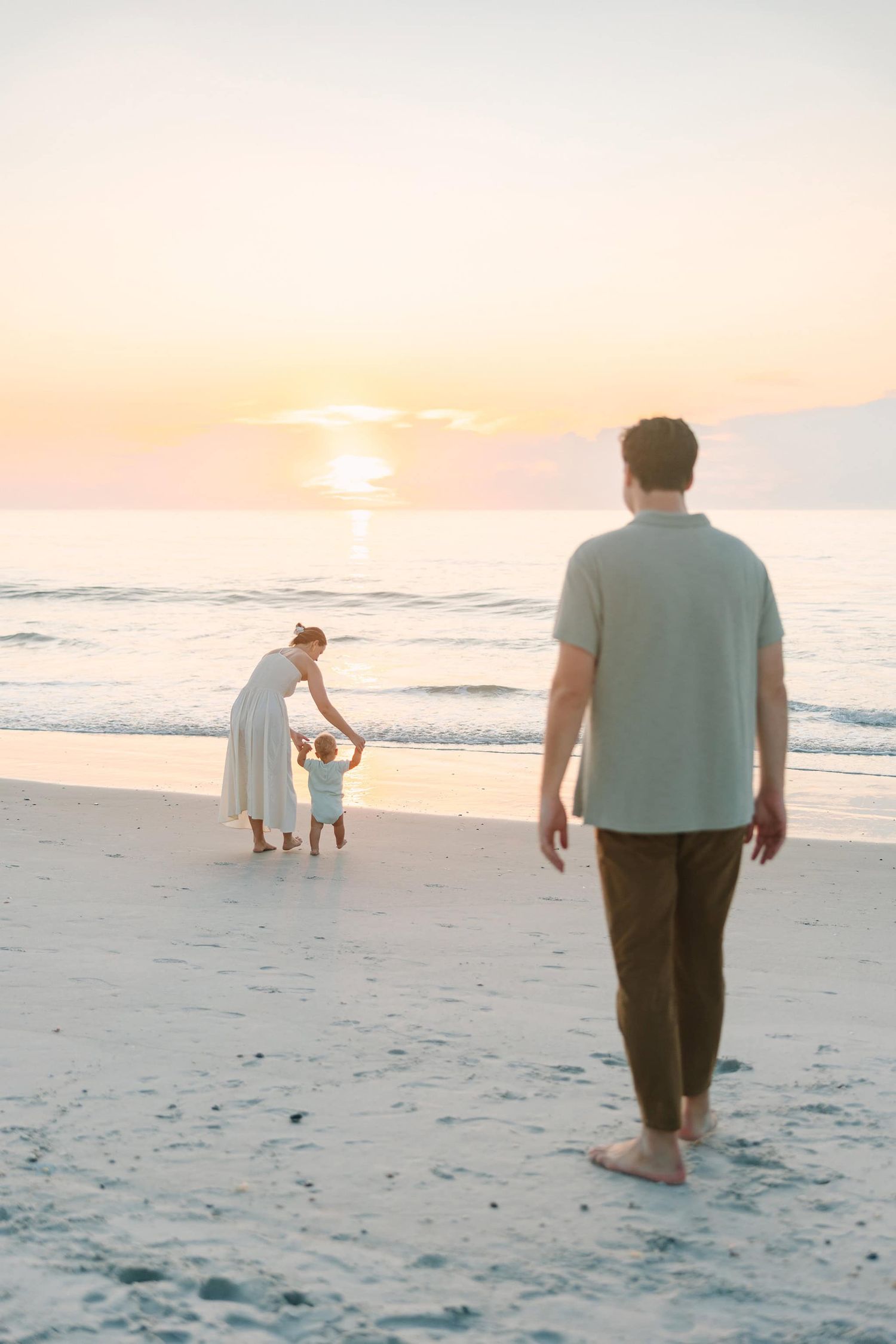 dad watching mom play with toddler son in the ocean as the sun rises over the ocean