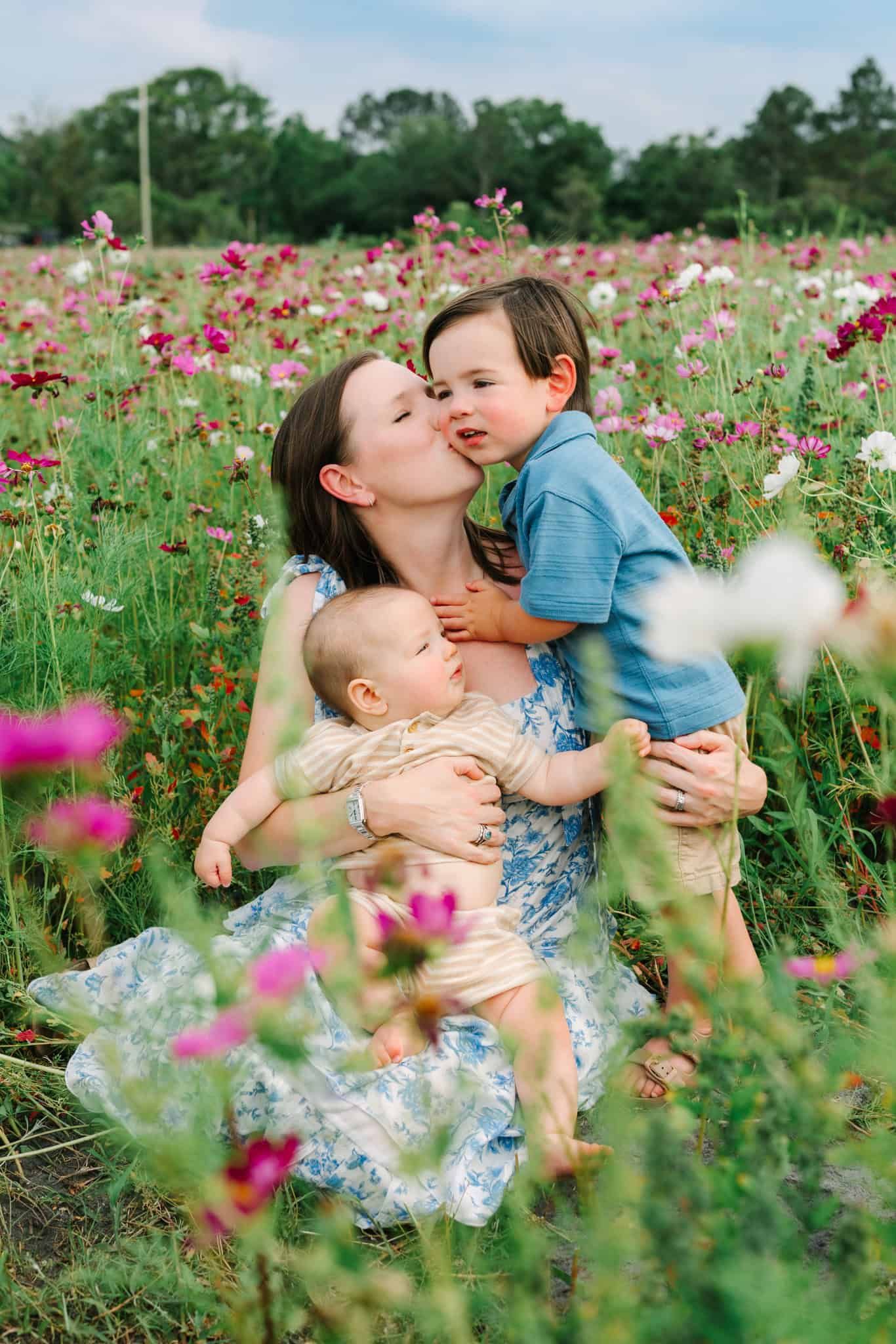 A mother kisses her toddler son while holding her baby in a vibrant flower field.