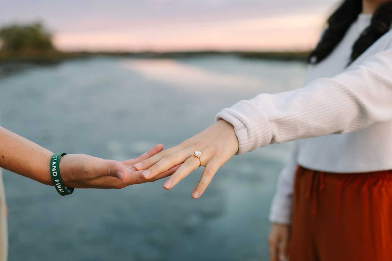 Newly engaged couple  touching hands outdoors near a lake at sunset, one wearing an engagement ring and a white sweater.