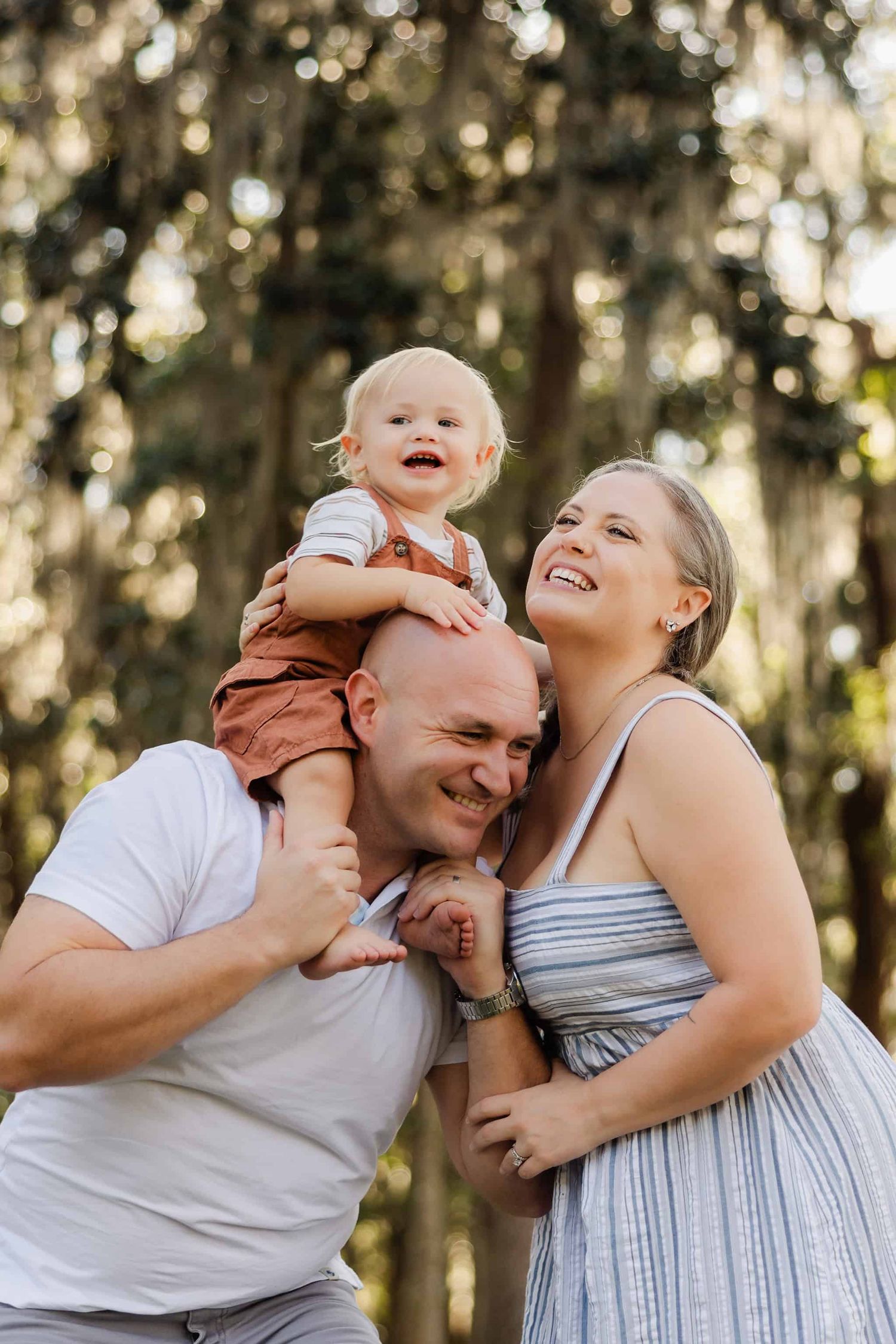 son on dads shoulders as dad leans for mom to give a kiss and everyone laughing. trees behind them in a Jacksonville park