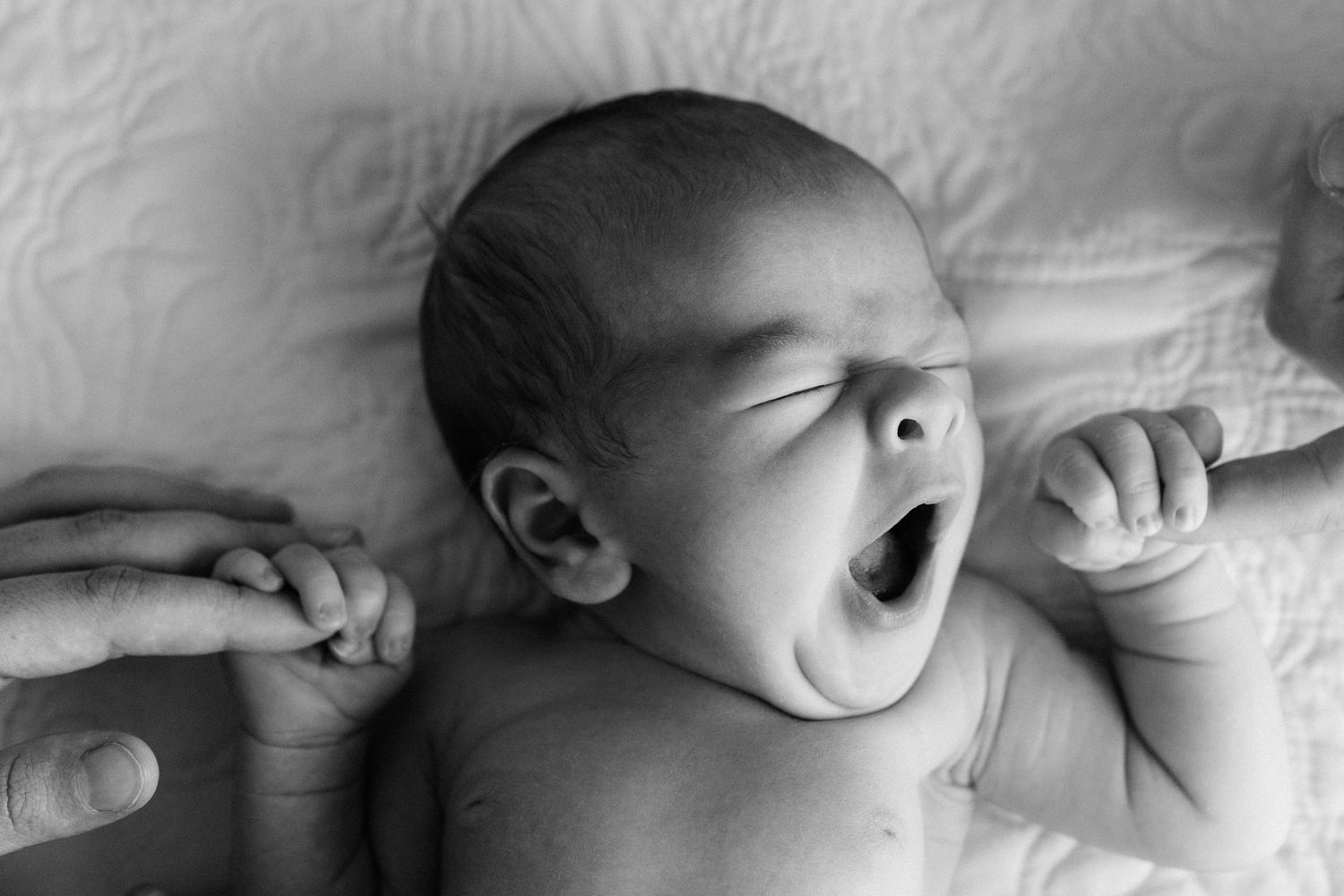 Black and white photo of a newborn baby yawning while lying on a white blanket, tiny hand reaching out.