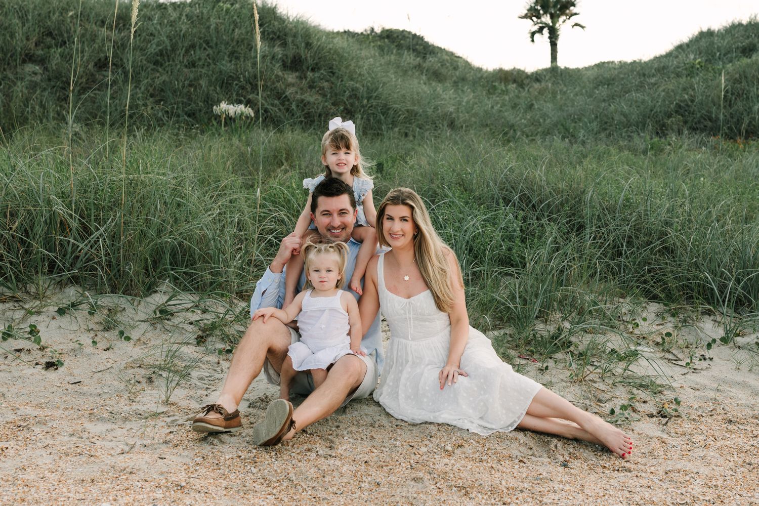 Happy family of four posing together on sandy Ponte vedra beach dunes with lush green grass backdrop during golden hour.