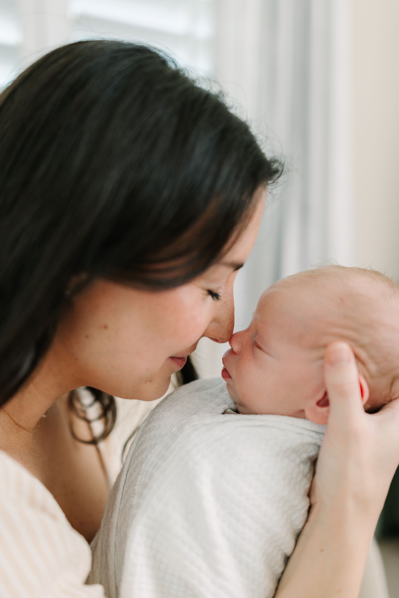 A mother tenderly touches noses with her swaddled newborn baby in a soft, intimate portrait.