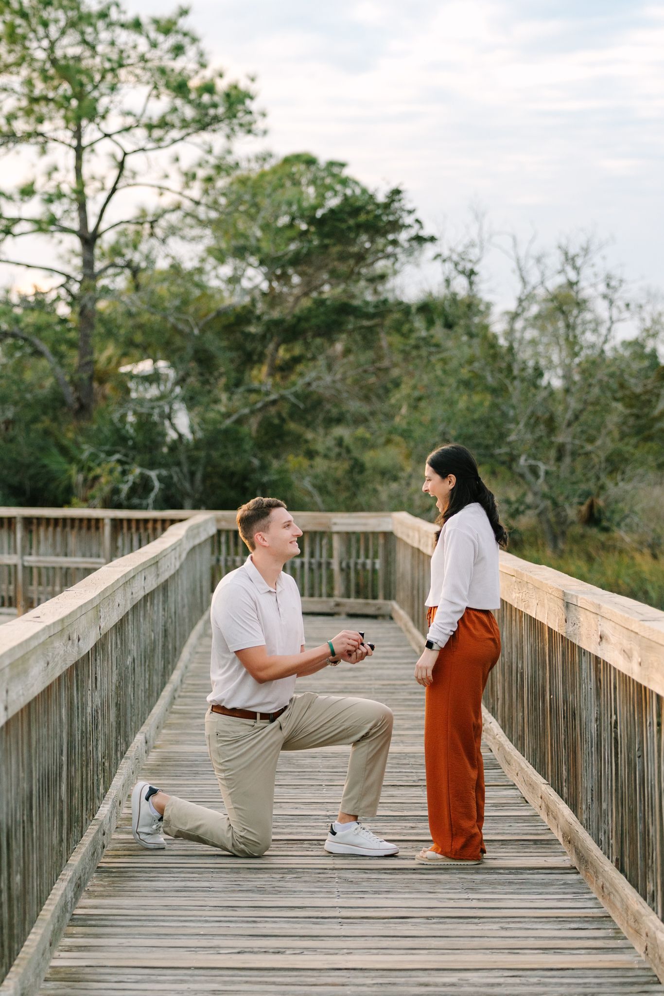Man proposing on one knee to woman on a wooden boardwalk surrounded by trees.