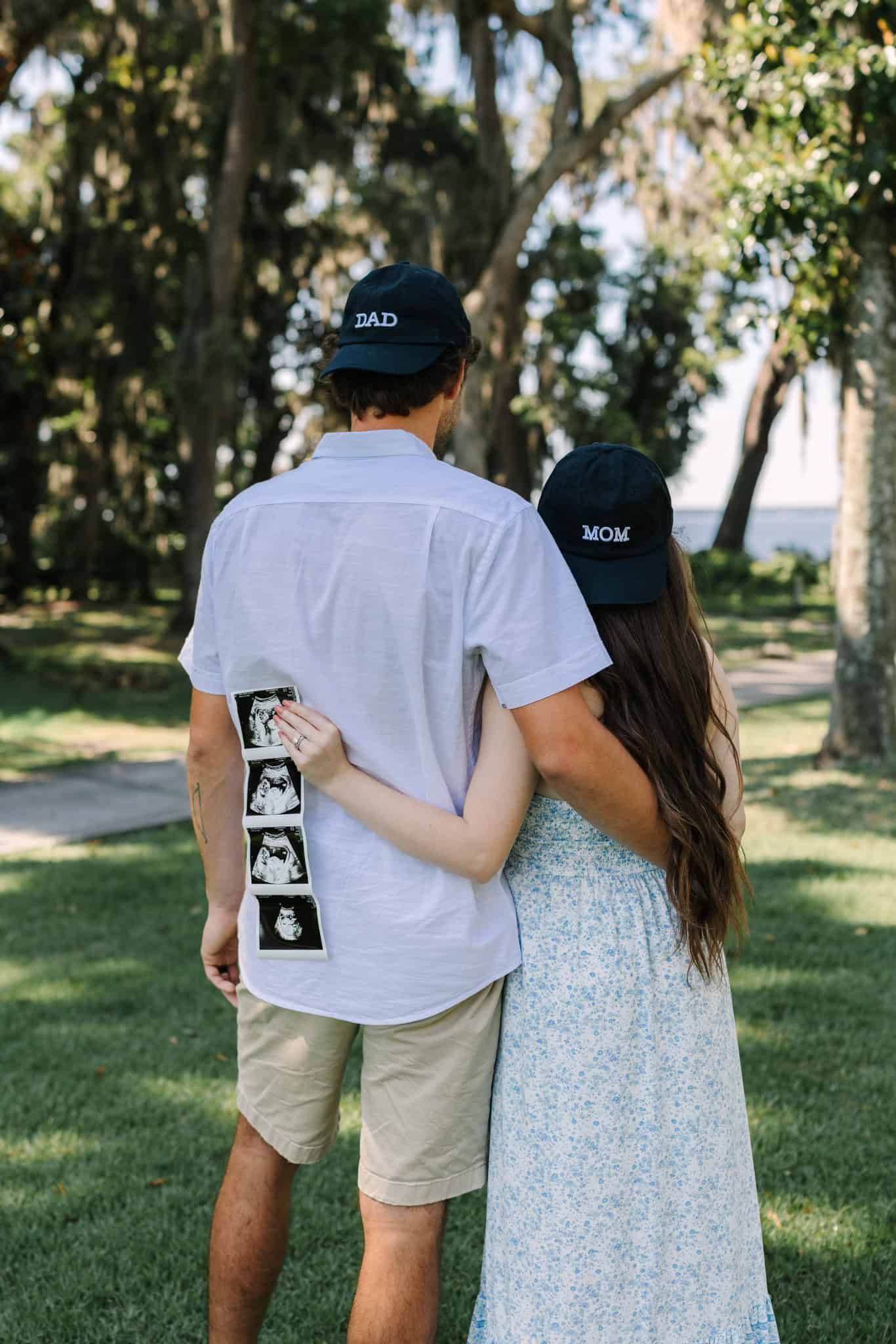 Couple wearing Dad and Mom hats from behind, holding ultrasound photos, announcing pregnancy outdoors.