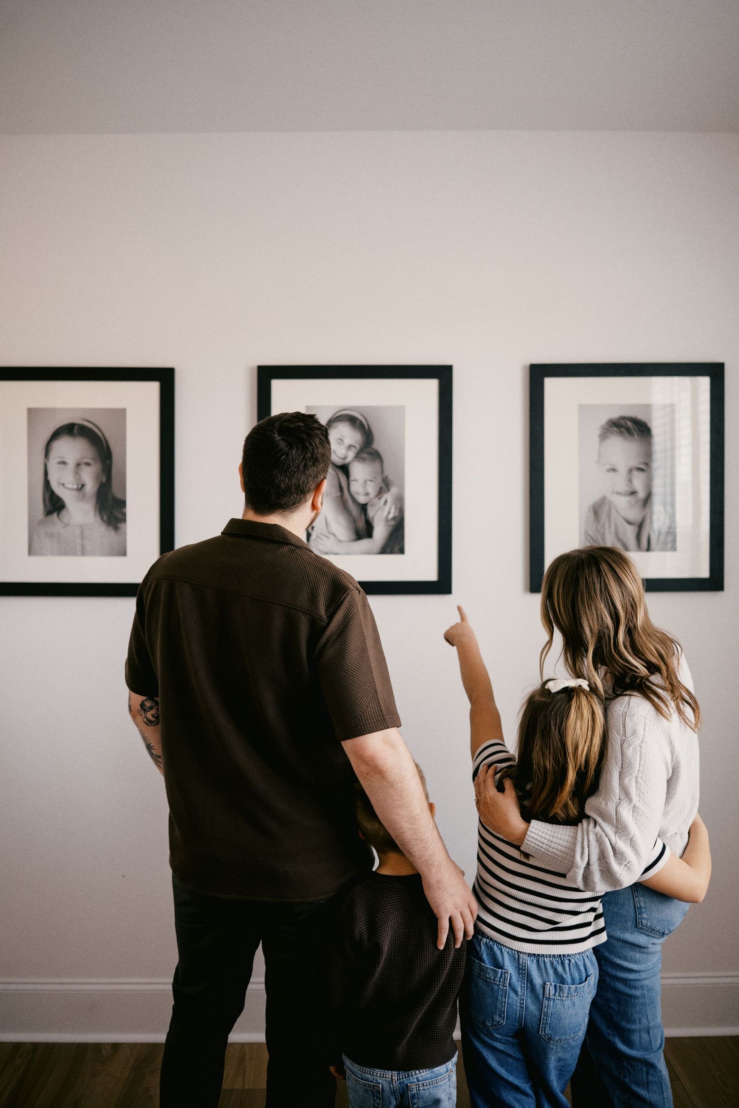 Family viewing black and white portrait photos on wall, mom pointing at framed children's pictures in home gallery.