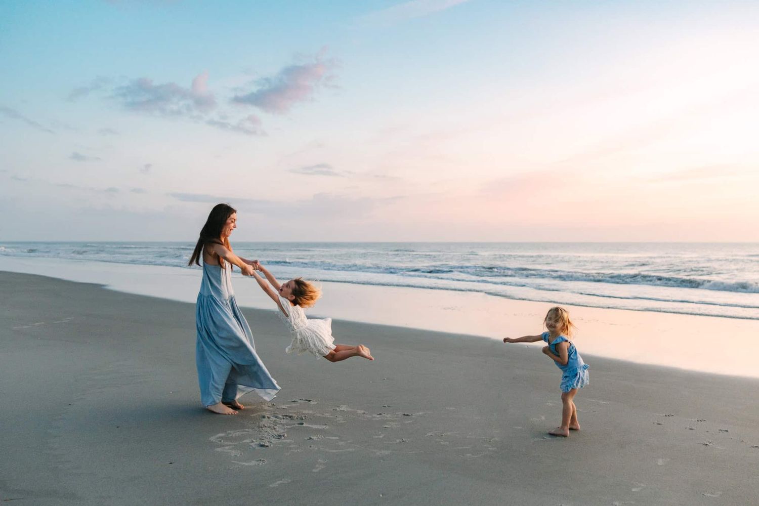 Mother in blue dress twirling children on sandy Ponte vedra beach at sunrise with ocean waves in background.