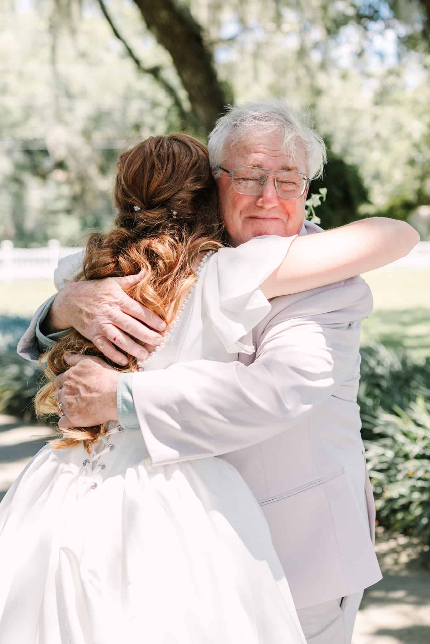 Emotional father-daughter hug at outdoor wedding, bride in white gown embracing older man in light suit among trees.