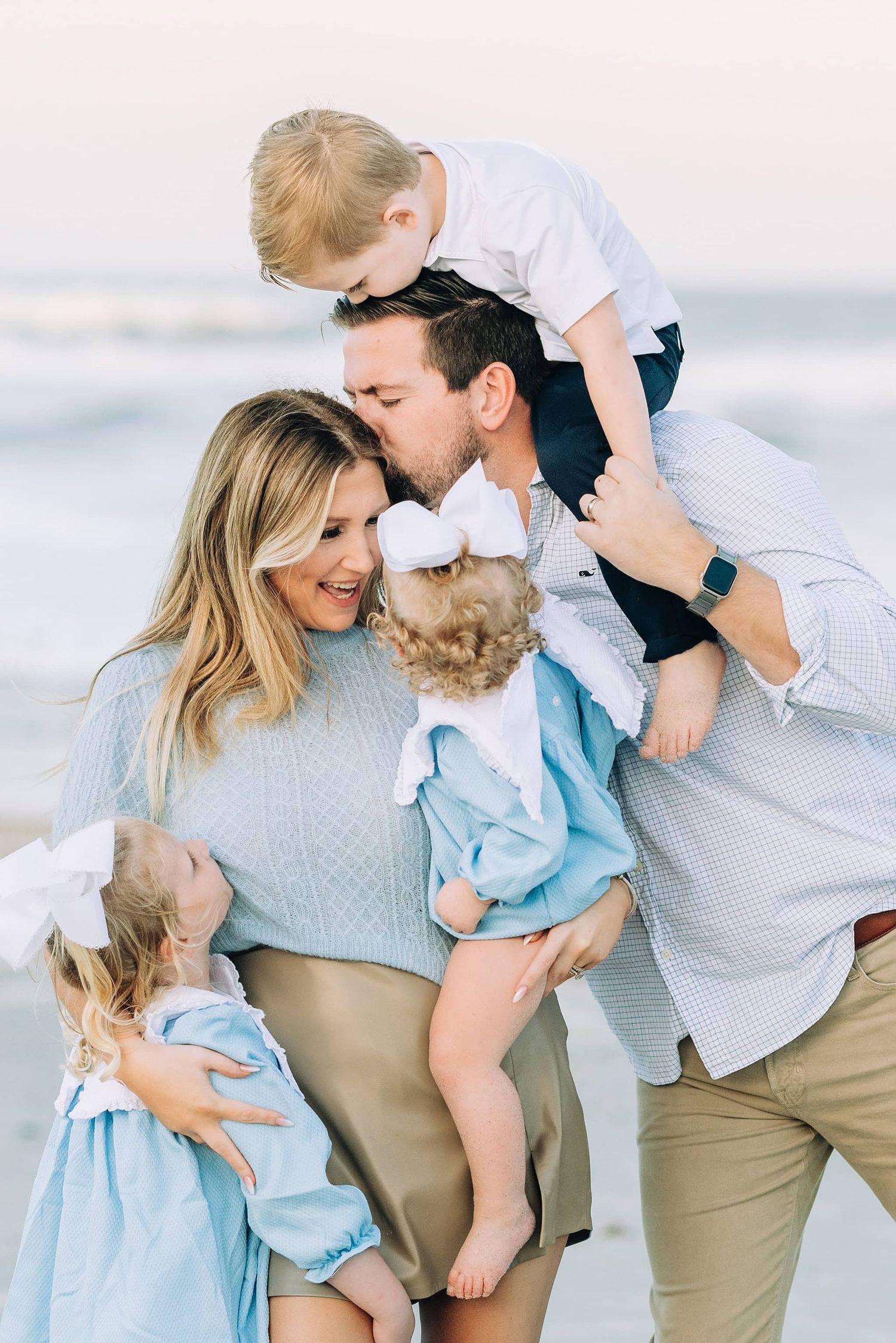 dad kissing mom on the temple with son on his shoulders and daughters giving mom big hugs at the beach with the water in the background