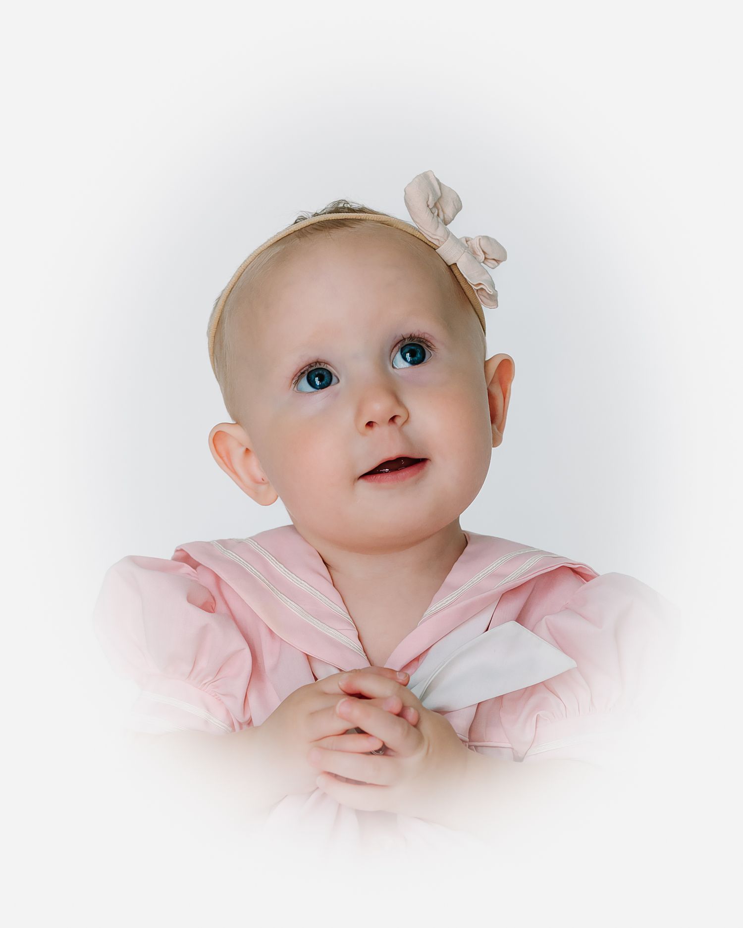 Adorable baby girl with blue eyes wearing a pink dress and bow headband, looking up with clasped hands.