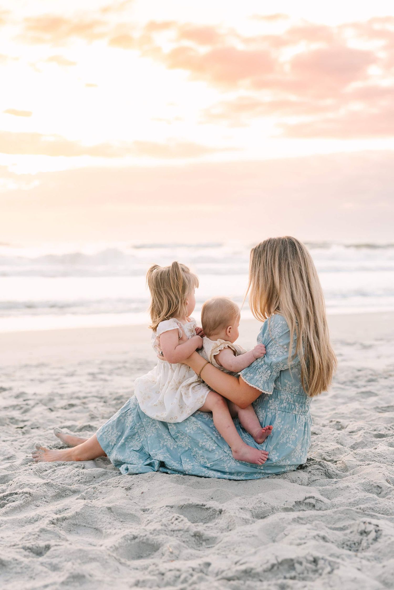 mom sitting in the sand with both daughters in her lap looking out at the sunrise over the ocean