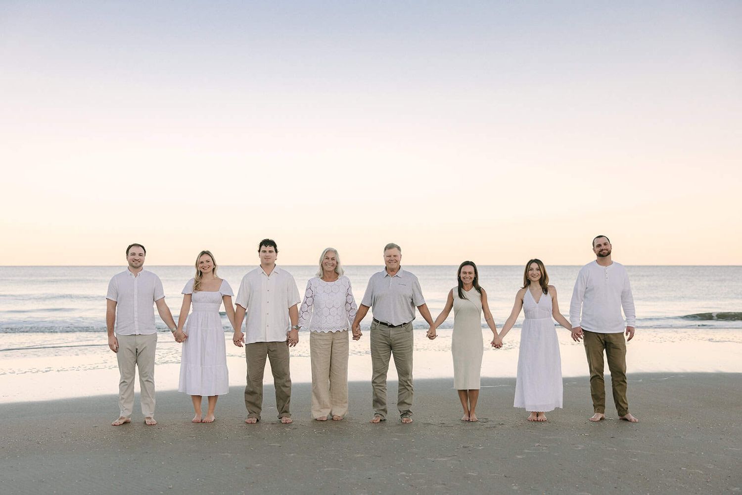 extended family holding hands standing on Jacksonville beach fl at sunset all wearing white