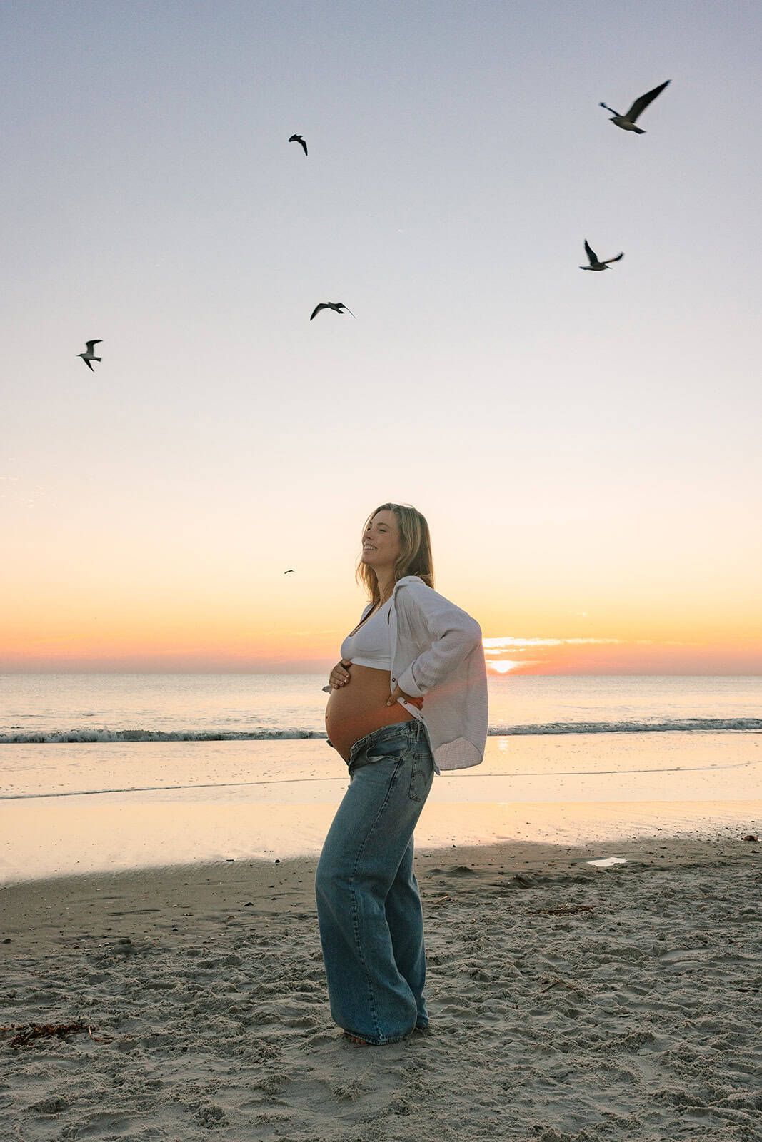 pregnant mama at Jacksonville beach sunrise looking up and laughing with her belly out and birds flying behind her