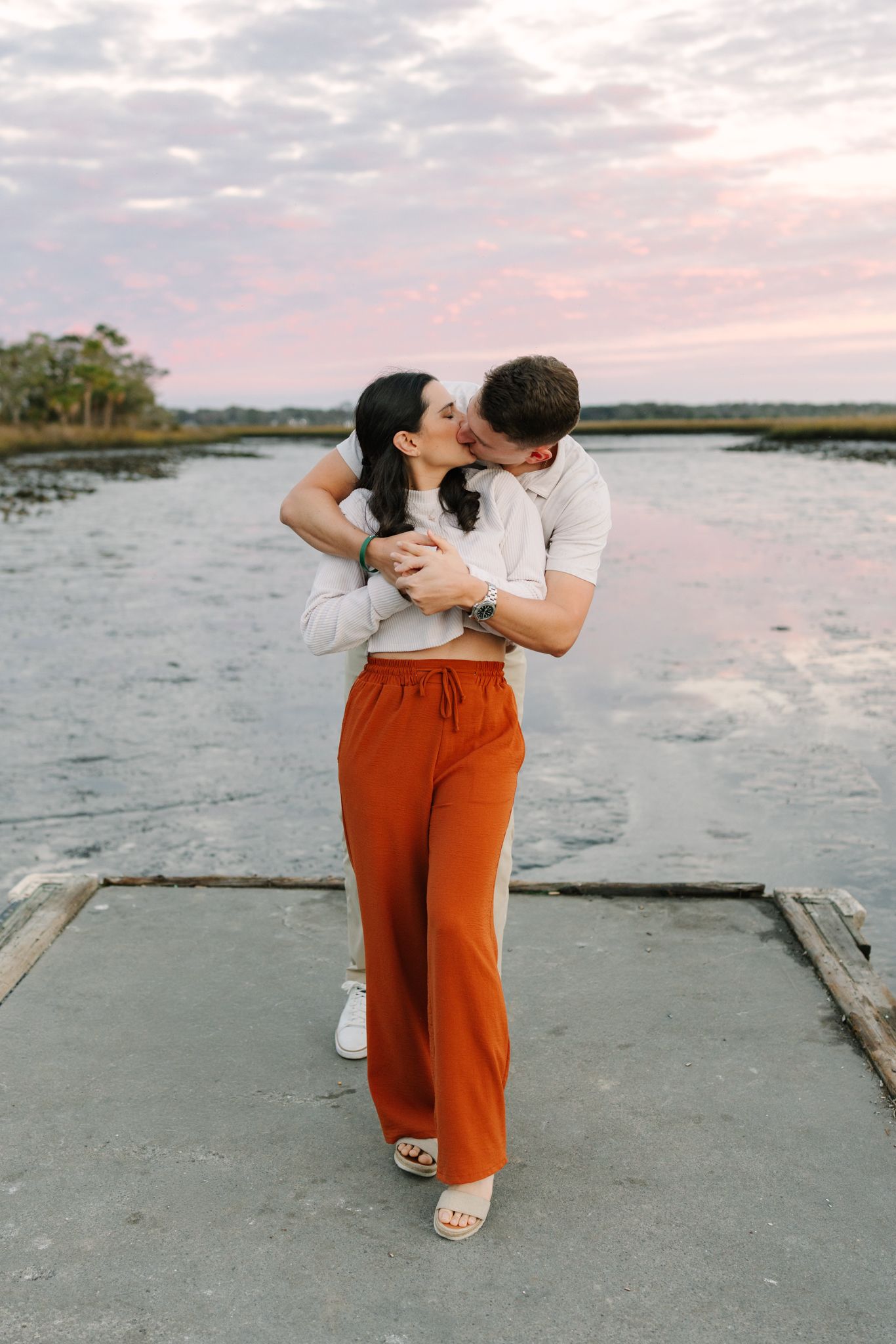 Couple sharing a romantic kiss on a dock at sunset, woman wearing orange wide-leg pants, marsh landscape behind them.