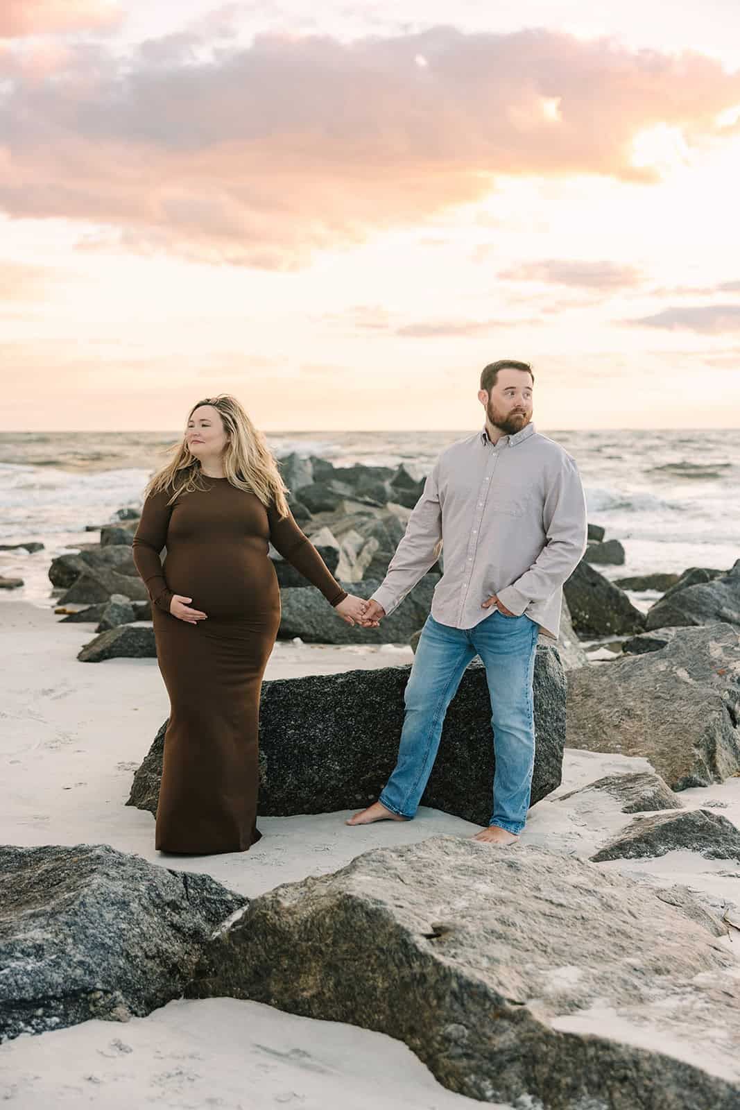 pregnant wife and husband holding hands in front of the jetty rocks and looking away. gorgeous sunrise of clouds and dimension