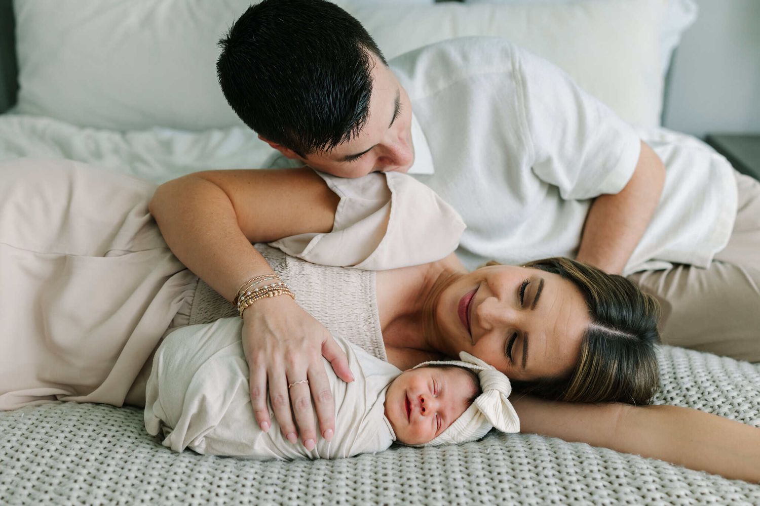 Parents lovingly cuddle their swaddled newborn baby on a bed in a tender family portrait.