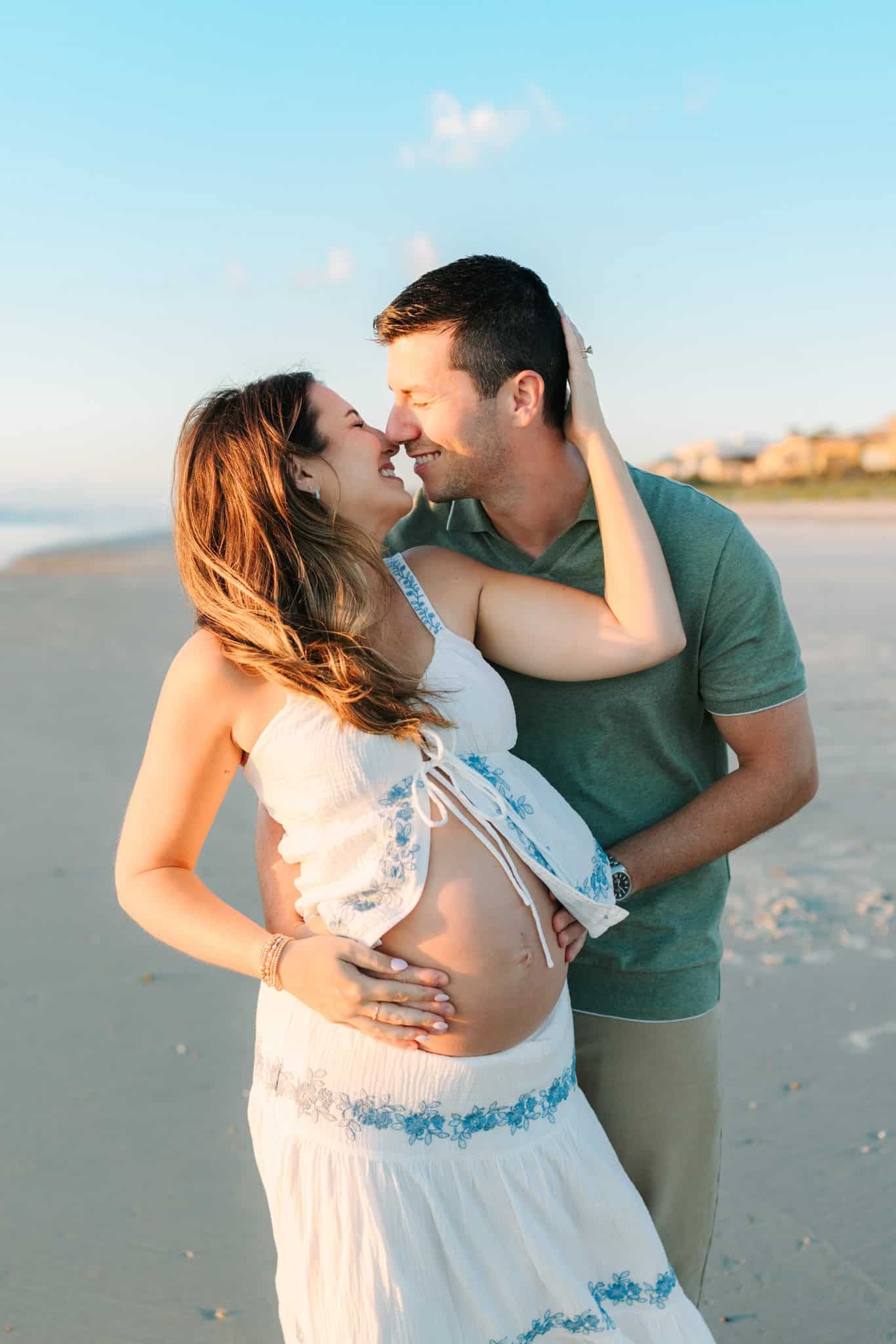 Pregnant couple embracing and smiling on a sunny beach, woman in white dress cradling baby bump.