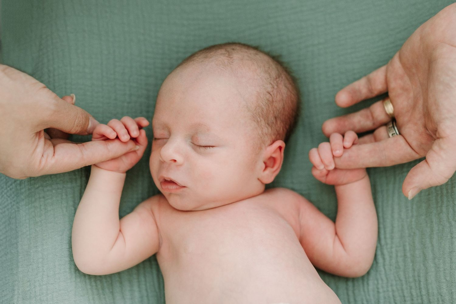 Newborn baby boy sleeping on green blanket while parents gently hold tiny hands in a tender family portrait