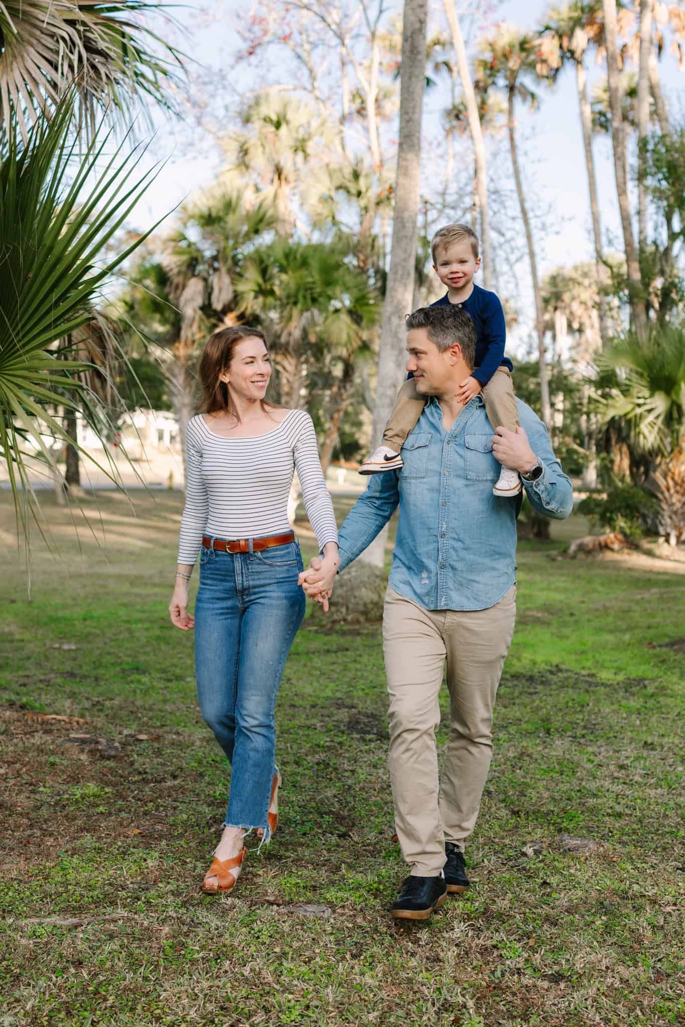 mom and dad walking through a park with lots of palm trees holding toddler boy, holding hands and smiling