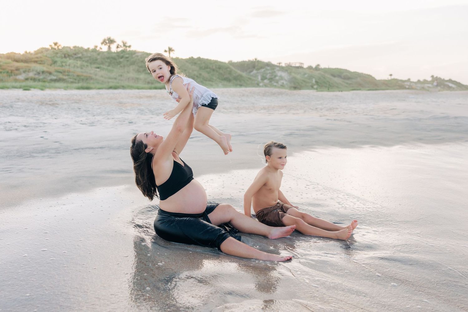 pregnant mama lifting her daughter in the air as she and her son sit in the water at the beach
