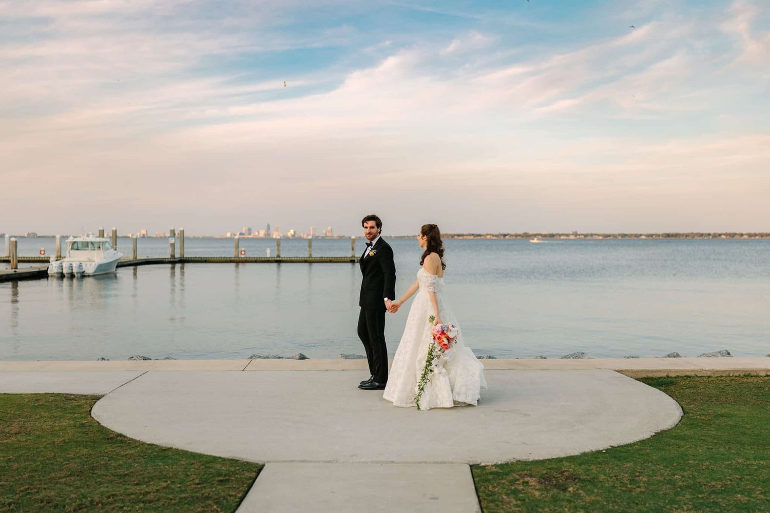 Bride and groom hold hands on waterfront path at sunset, with dock and calm bay in background.