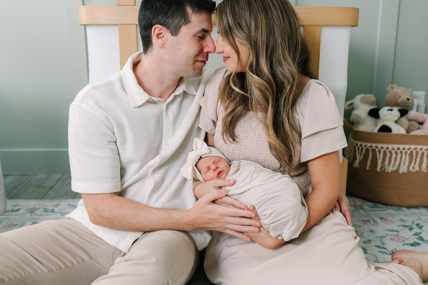 New parents sitting on nursery floor gazing lovingly at their swaddled newborn baby in a softly decorated room.
