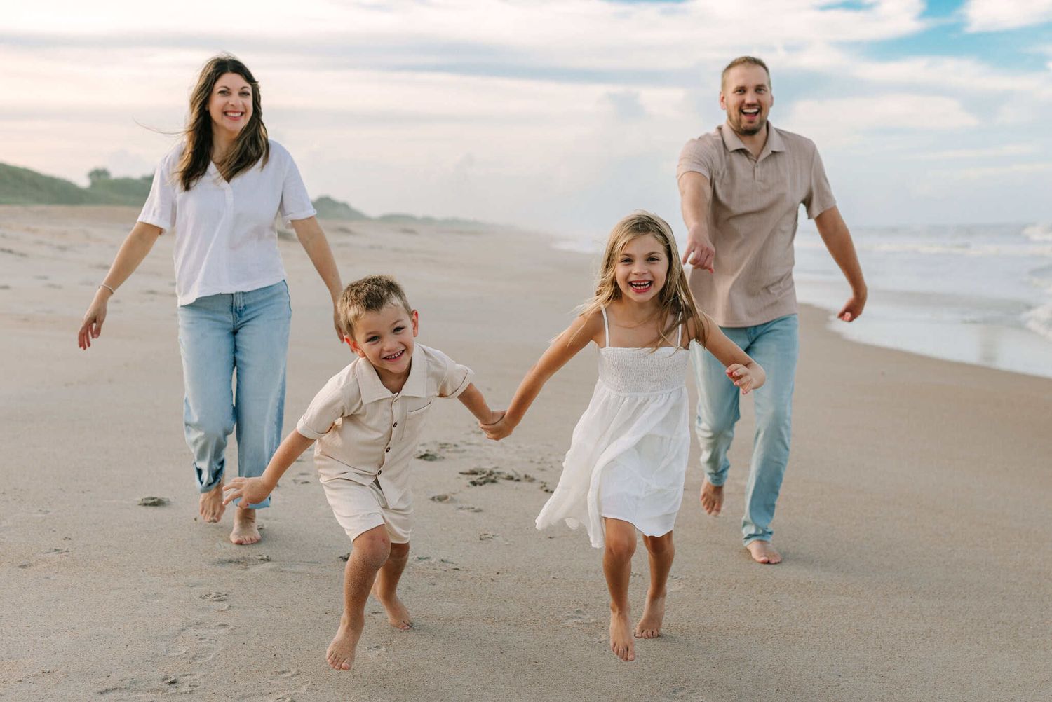 Happy family of four running and laughing on a sandy beach at sunset, parents and two young children holding hands.