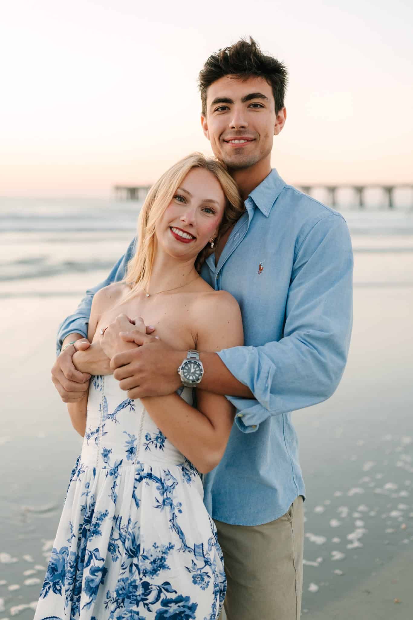 A smiling couple embraces on jacksonville beach at sunrise, woman in floral dress, man in light blue shirt wearing a watch.