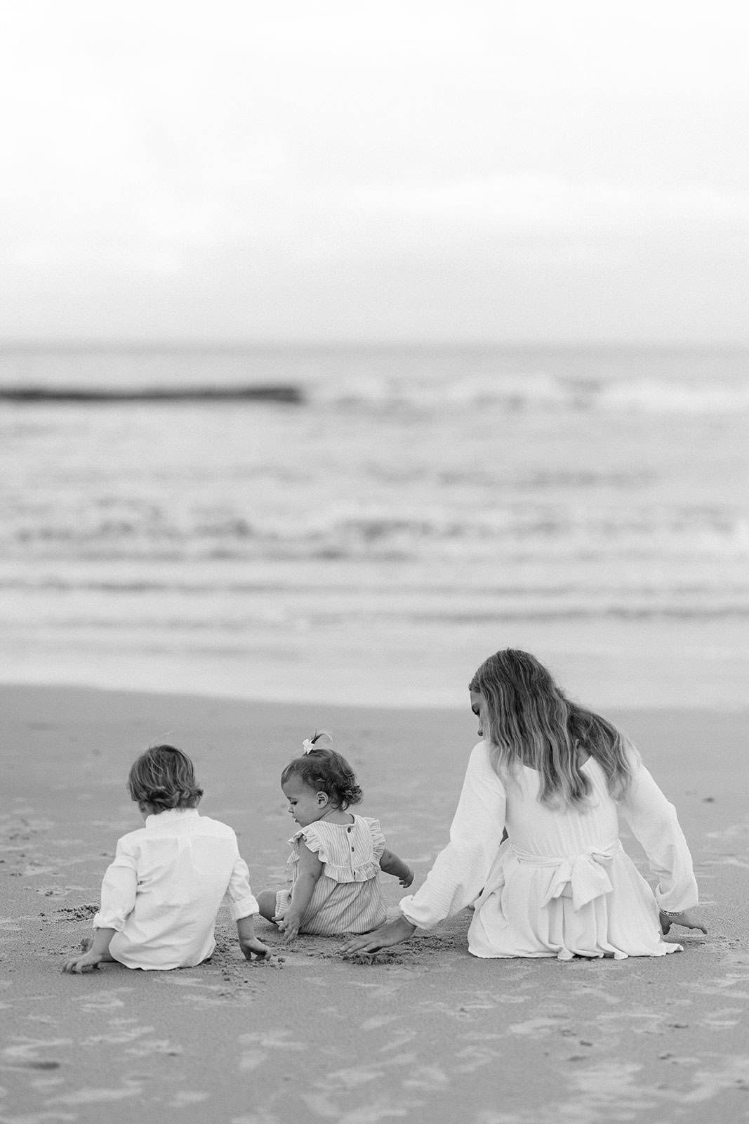 black and white photo of kids sitting playing in the sand and looking out at the ocean
