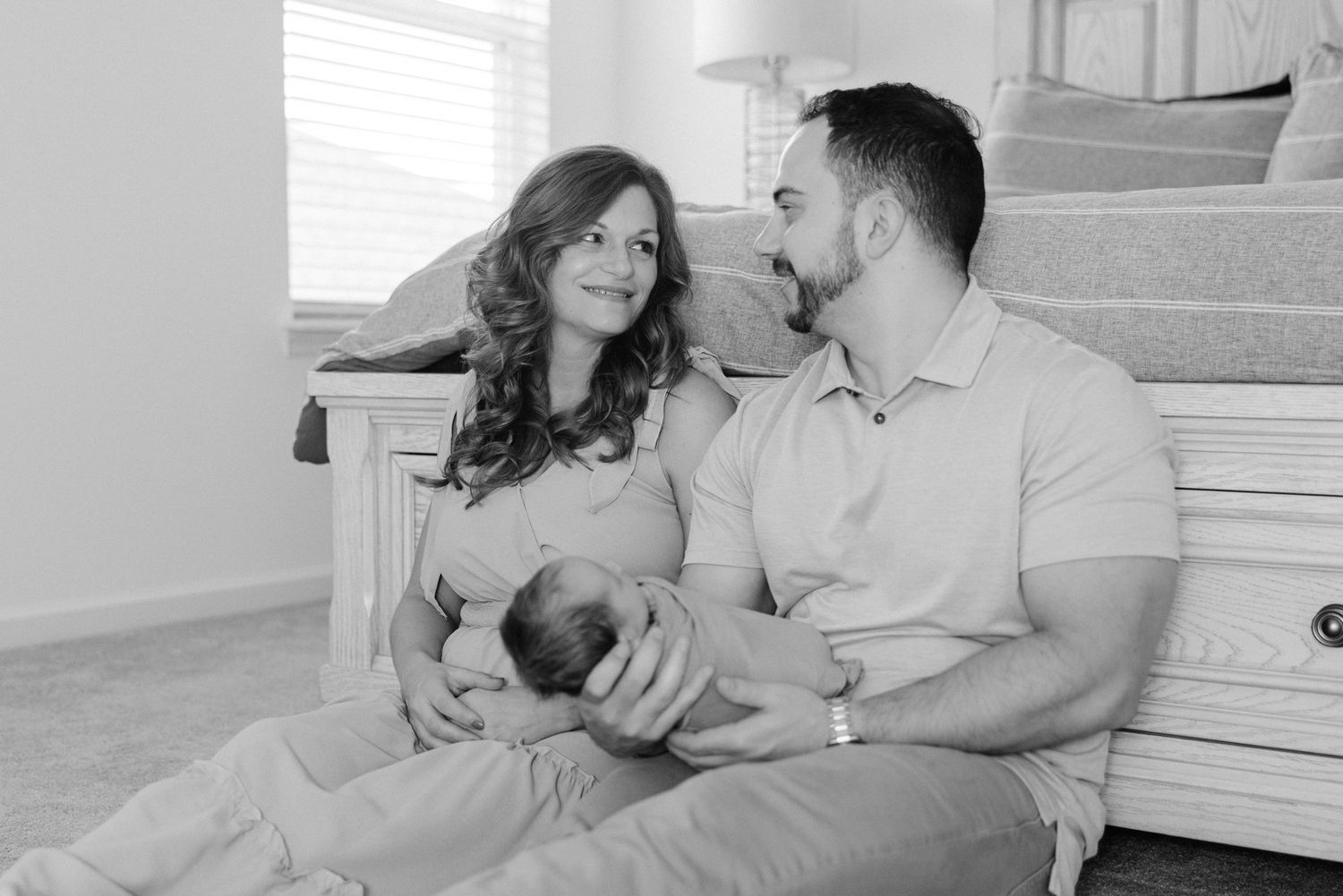 black and white image of parents holding newborn baby sitting at the foot of their bed