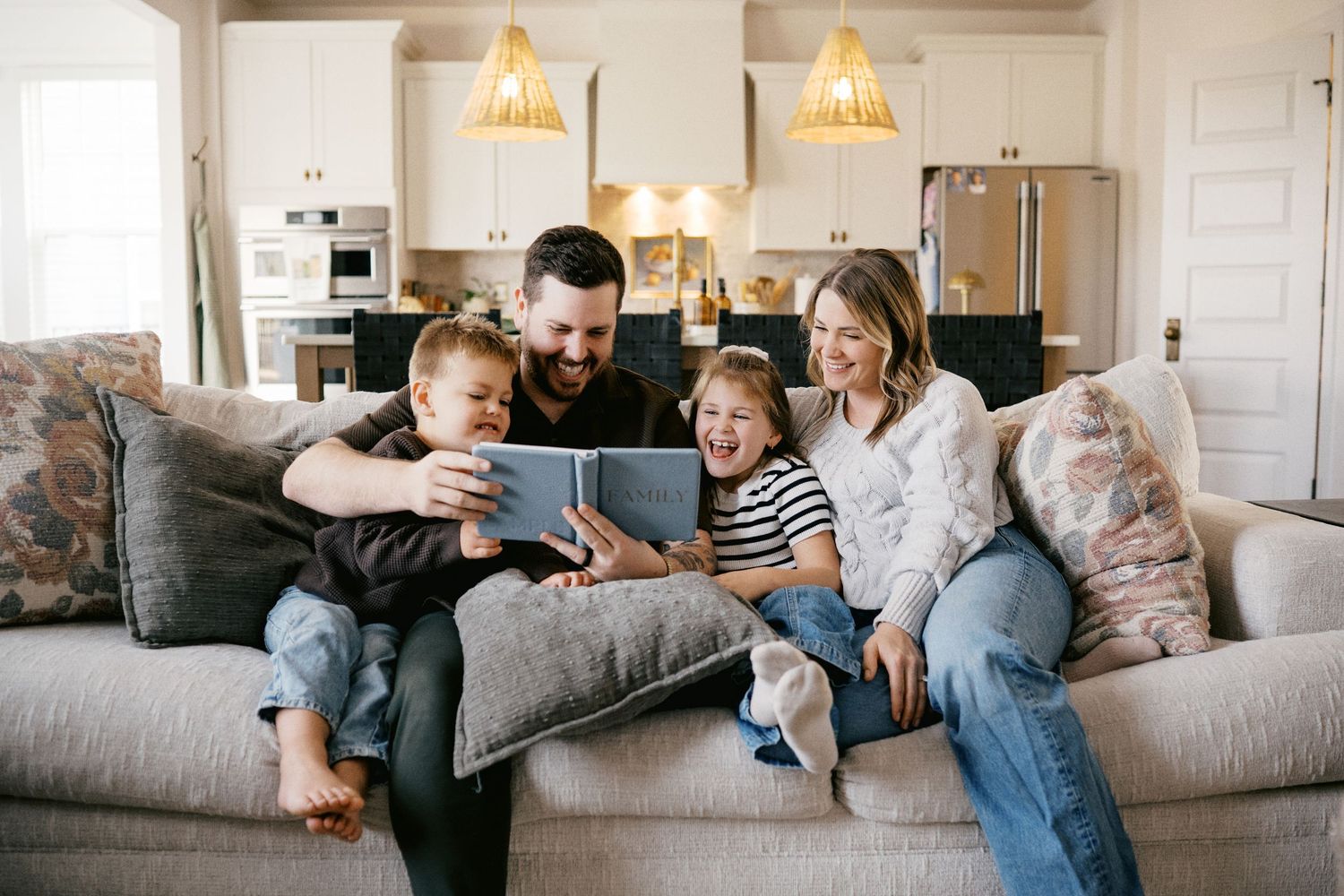 Family of four laughing together on a couch while looking at a tablet in a bright modern home.