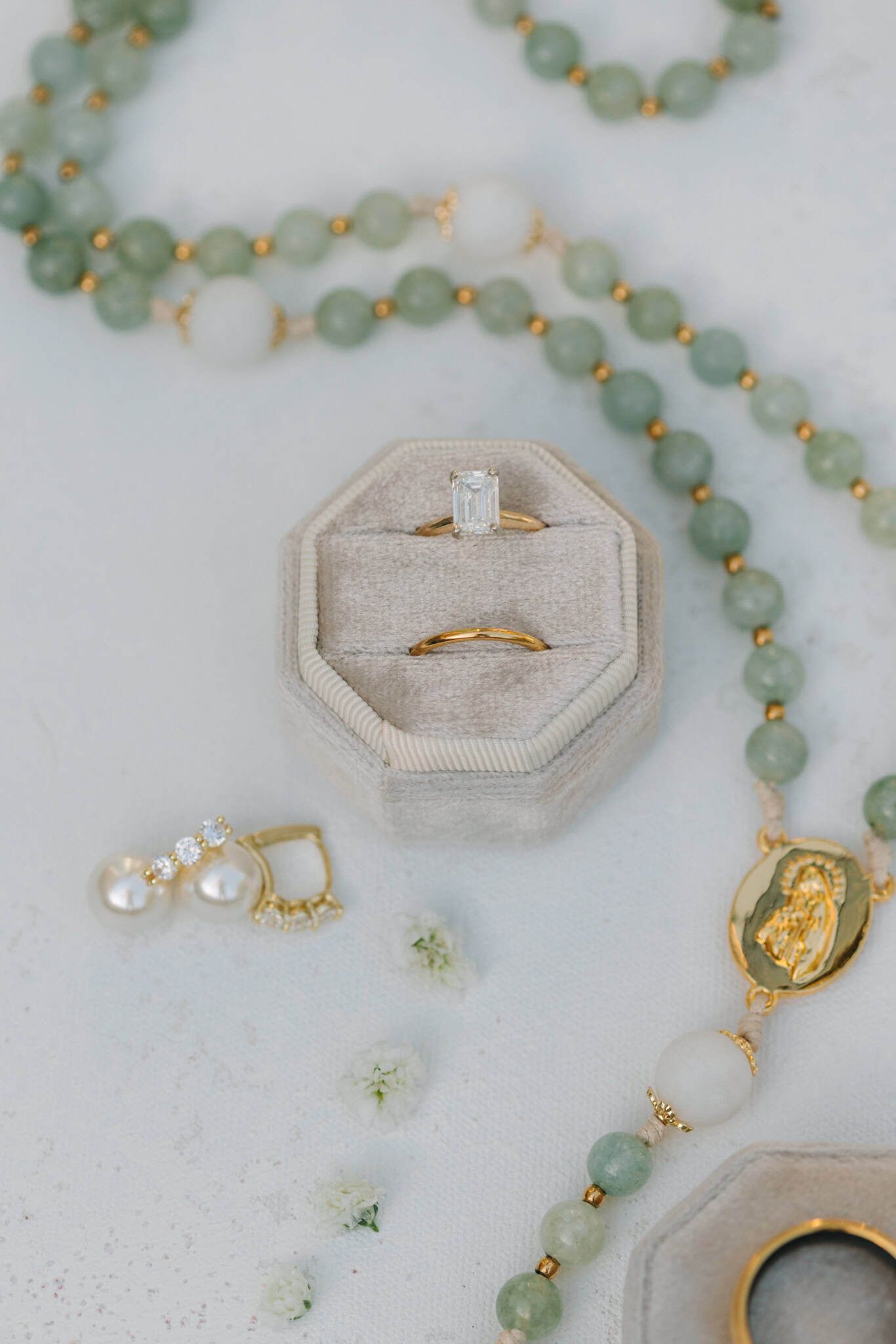 Gold engagement ring with emerald-cut diamond in velvet box, pearl earrings, and green jade beaded rosary on marble.