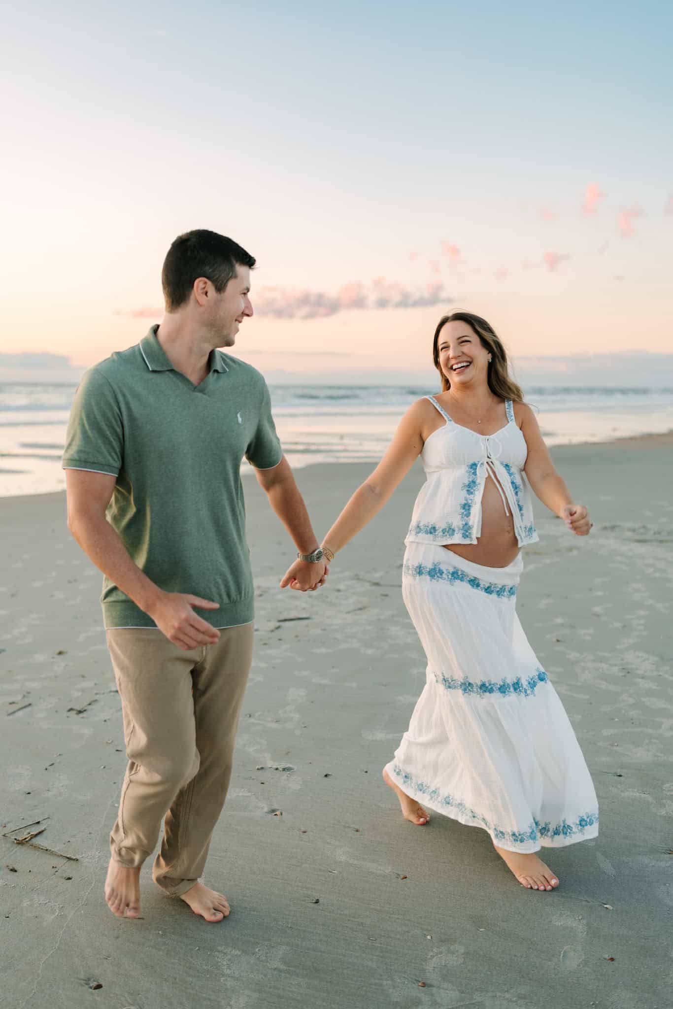 Couple holding hands and laughing on a beach at sunset, woman in white embroidered dress showing baby bump.