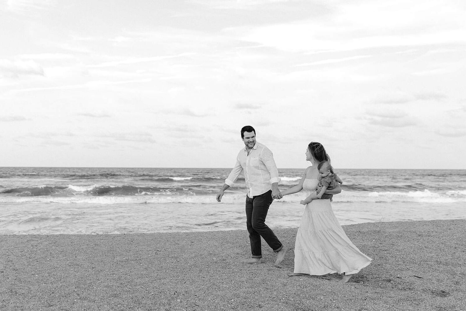 black and white photo of dad leading mom down the beach with her dress flowing while she holds their son