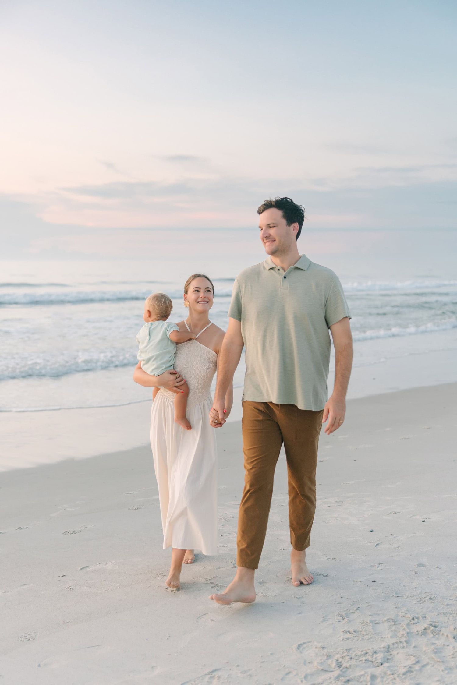 mom and dad holding hands walking down Jacksonville Beach during sunrise while mom holds son
