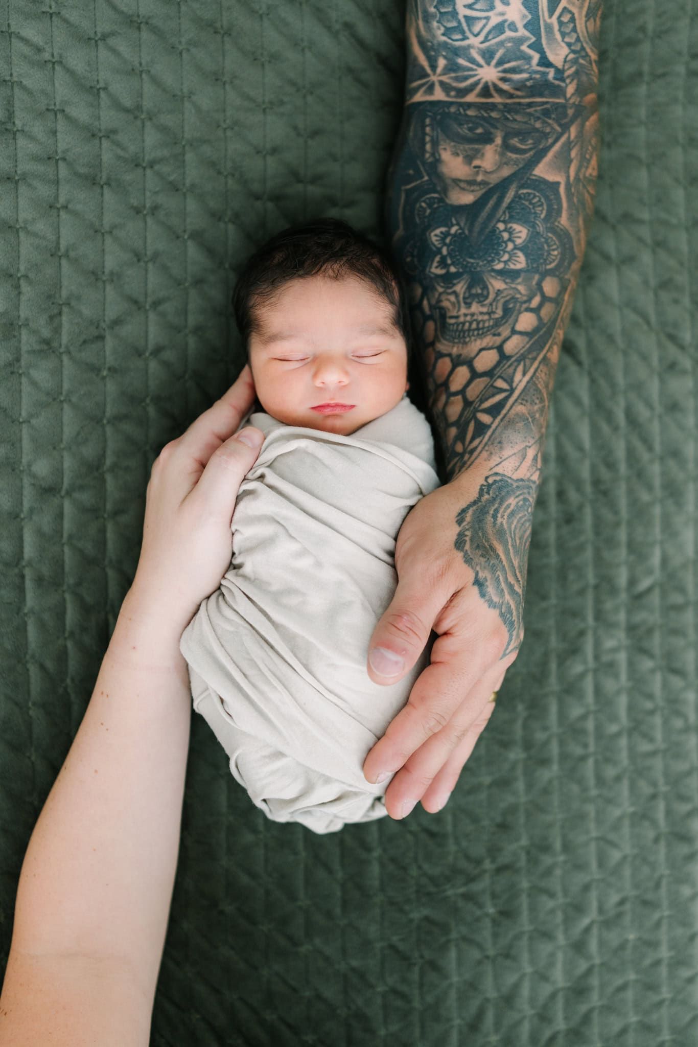 Newborn baby swaddled in white, cradled by two hands, one with tattoos, on a green quilted blanket.
