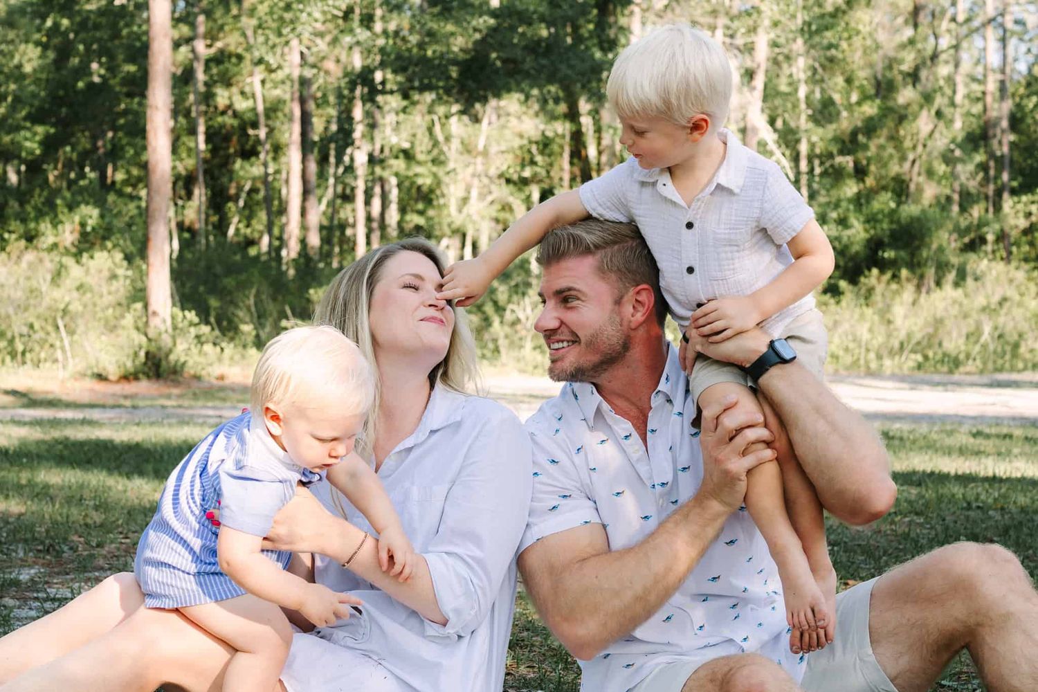Happy family of four sitting outdoors on grass, parents smiling with two young blonde boys in a sunny park setting.
