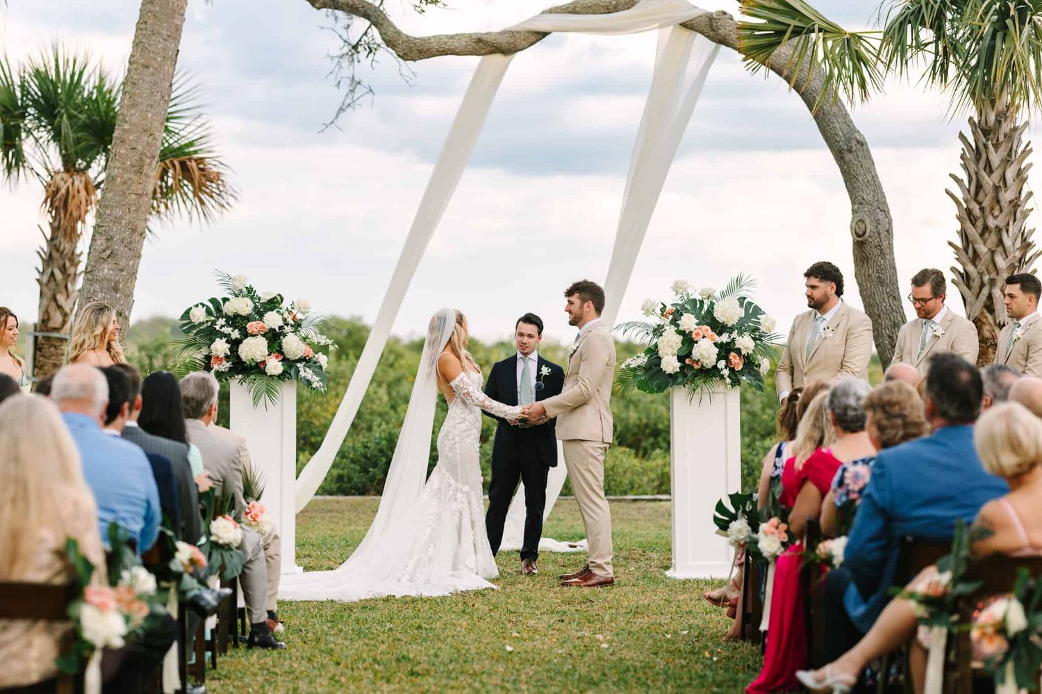 bride and groom holding hands smiling at each other at the altar, outside under a tree with drapery