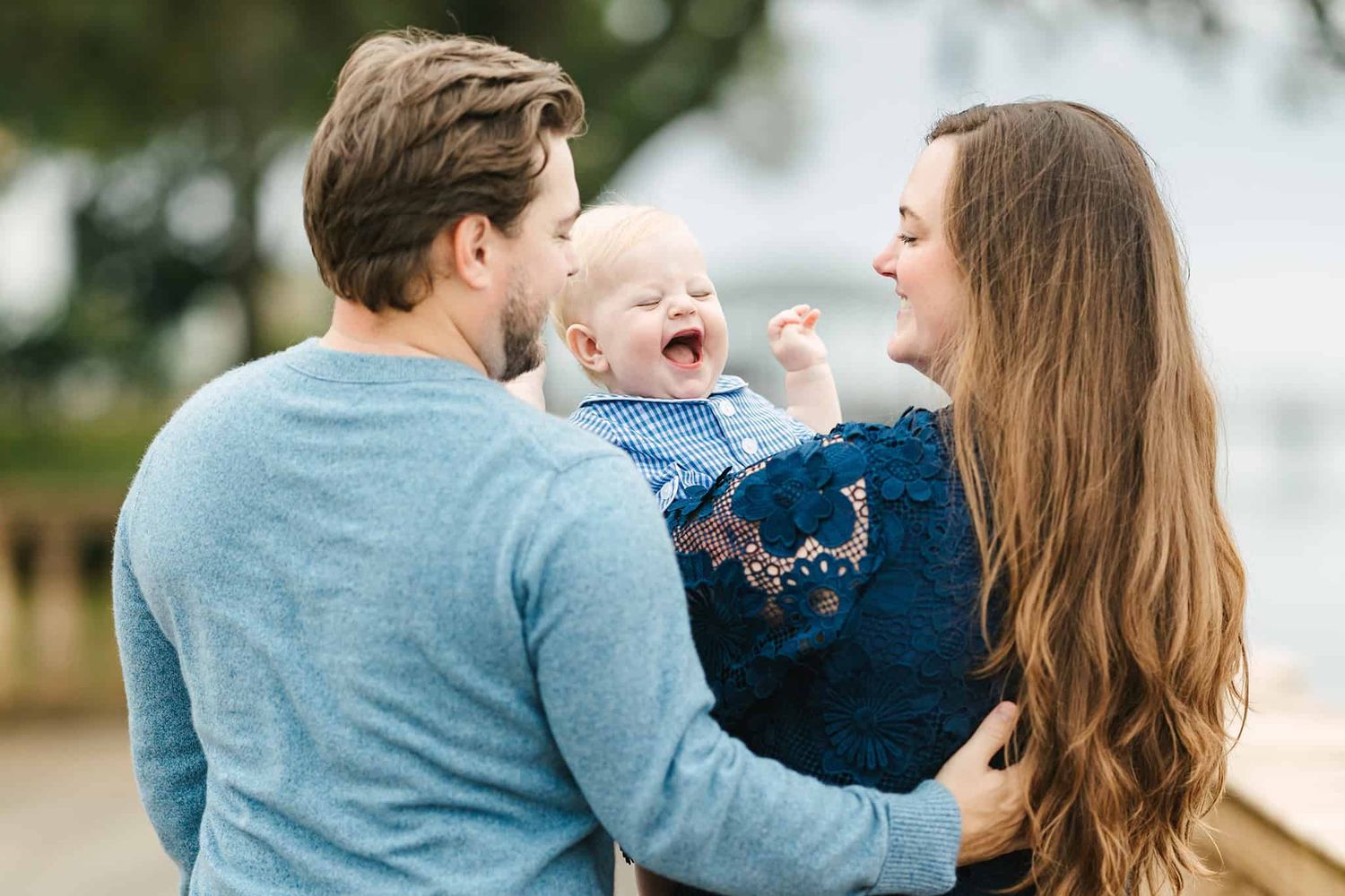 baby boy with huge smile and laughing and peeking over mom and dad who are facing away from the camera
