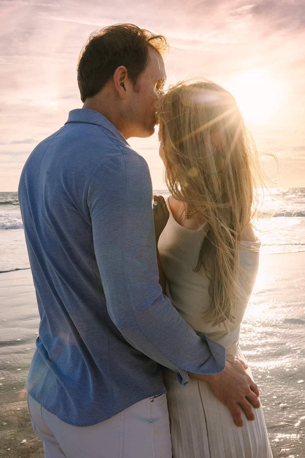 Couple sharing a romantic kiss on the beach at golden sunrise, waves in background.